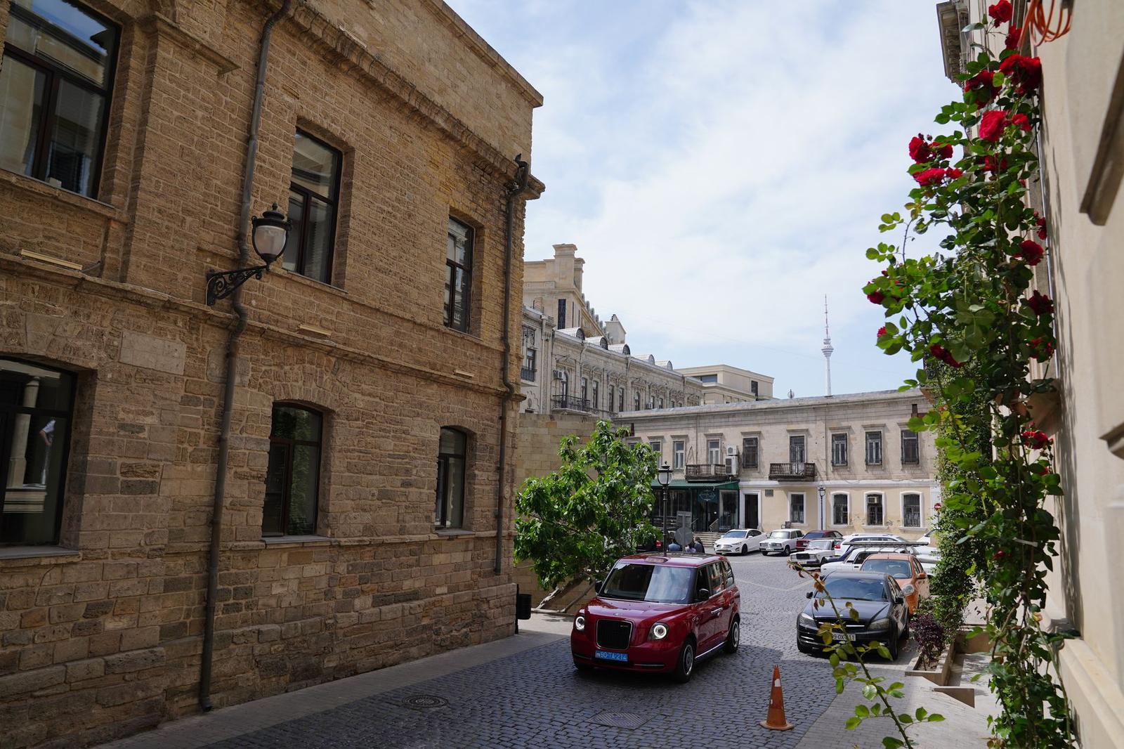 A red car parked on a cobblestone street beside stone buildings and climbing roses