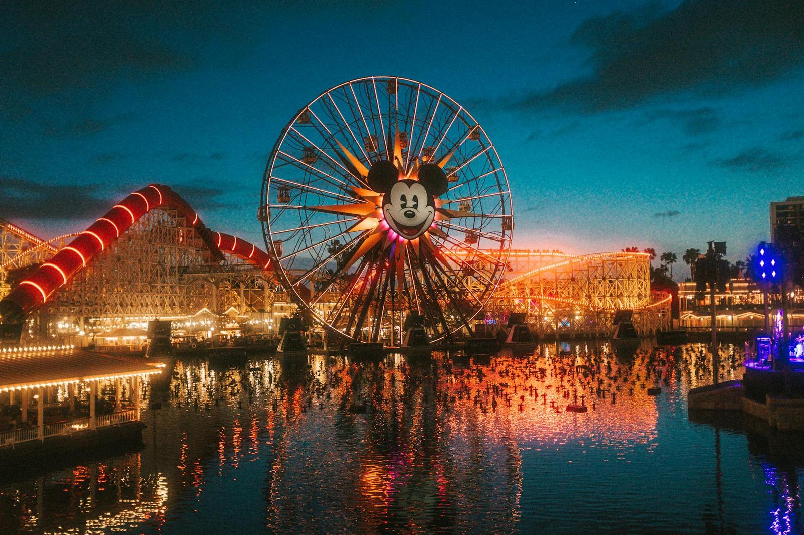 Illuminated ferris wheel with a Mickey Mouse design reflected on water at dusk