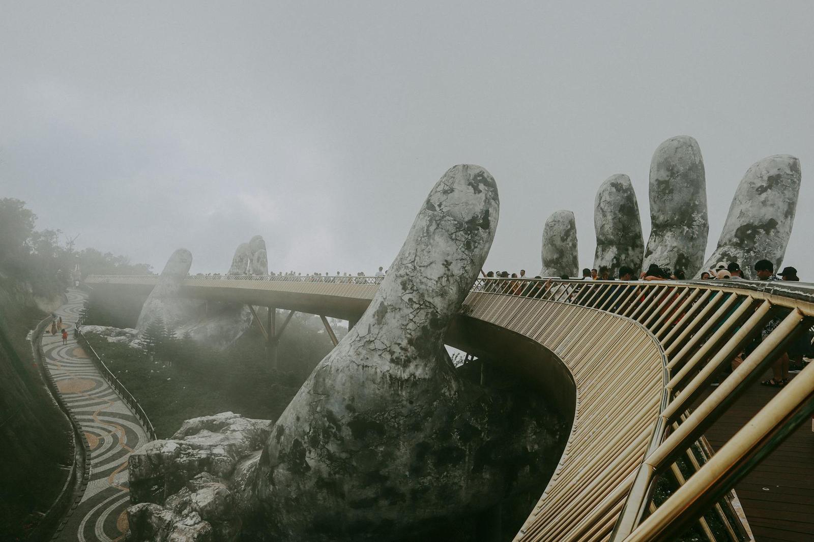 Visitors crossing a golden footbridge cradled by giant stone hands in thick fog