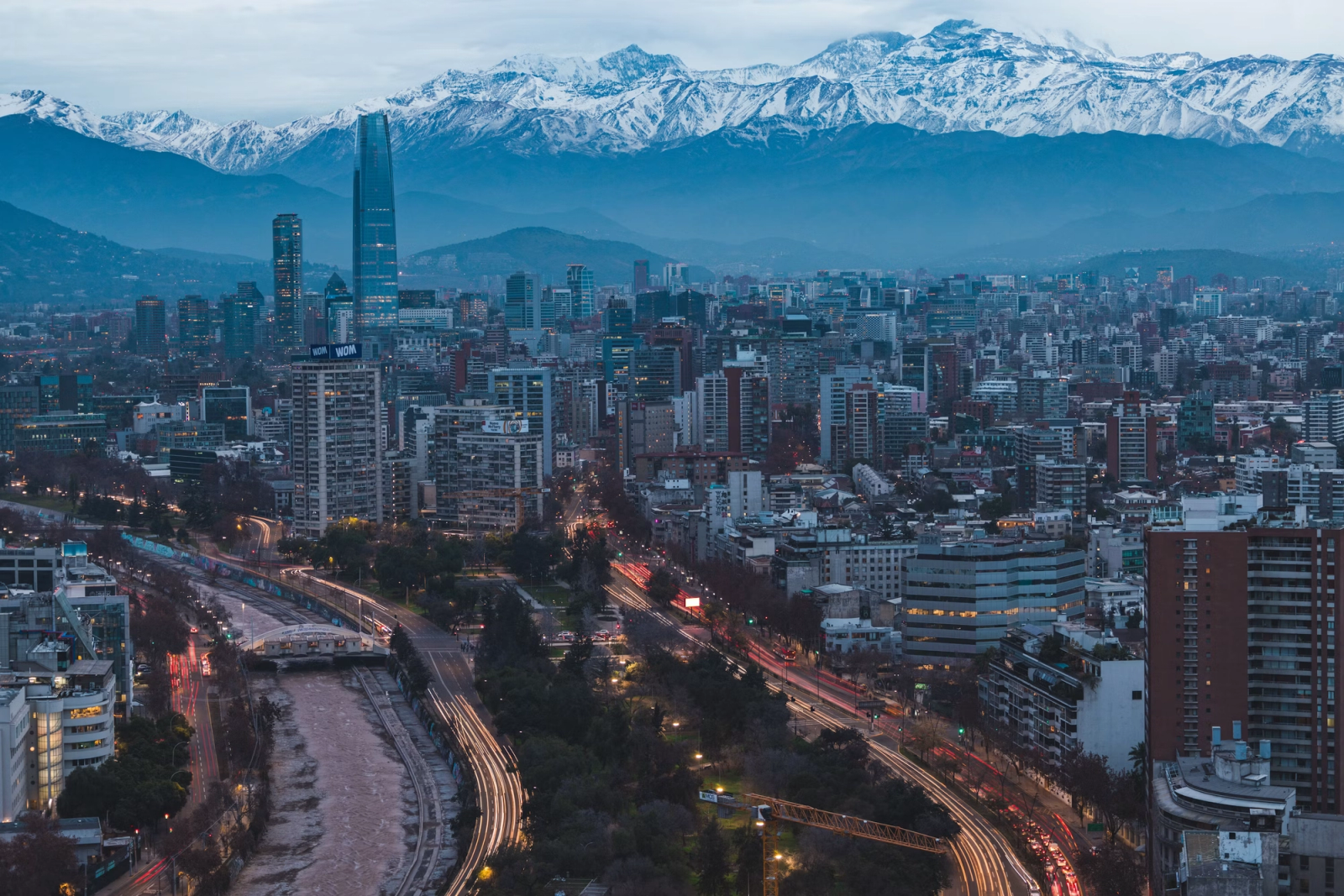 Snow-capped mountains loom behind a dense city skyline at dusk with light-trailed roads