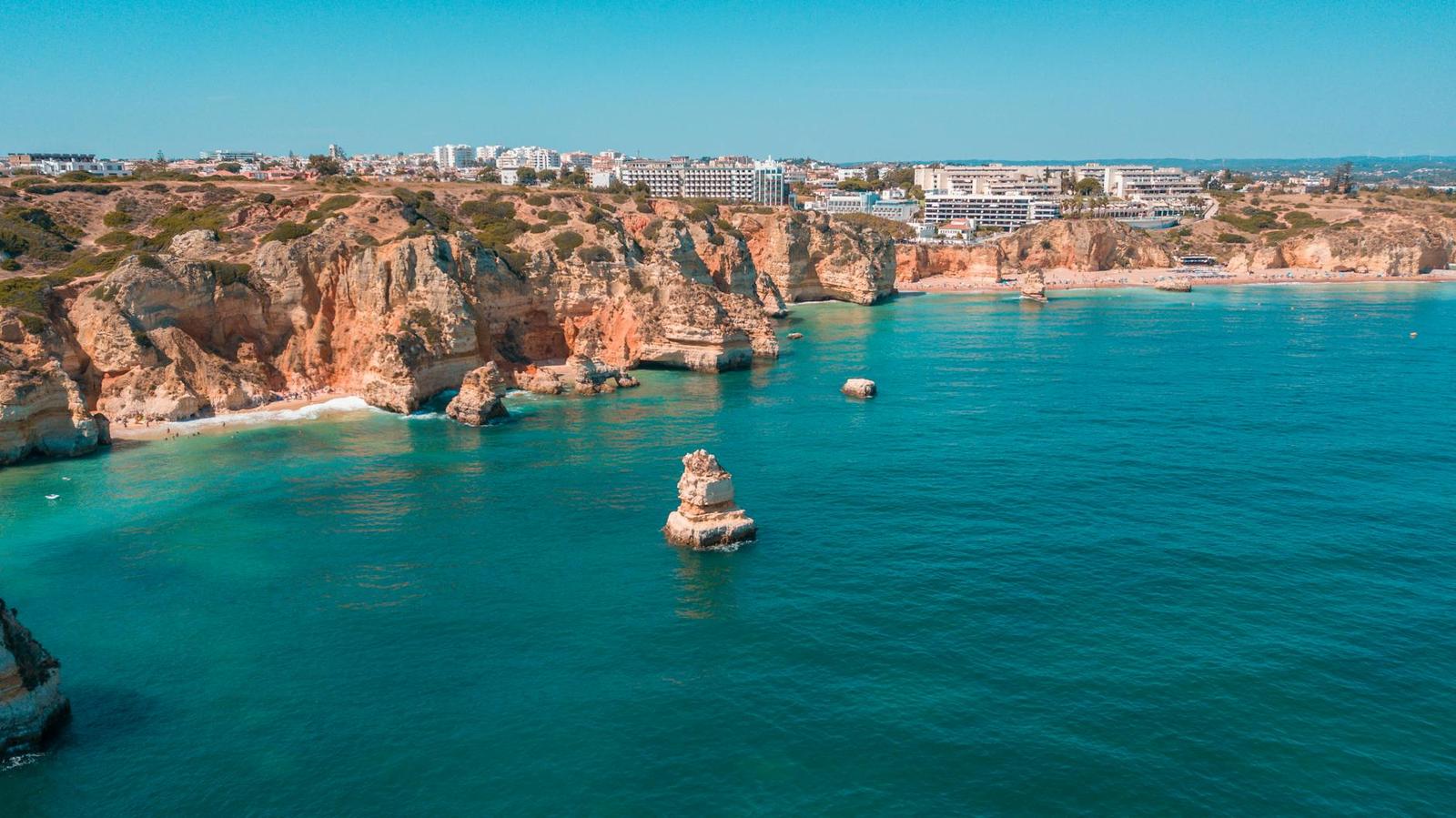 Aerial view of orange sea cliffs and a rock stack beside a resort-lined bay