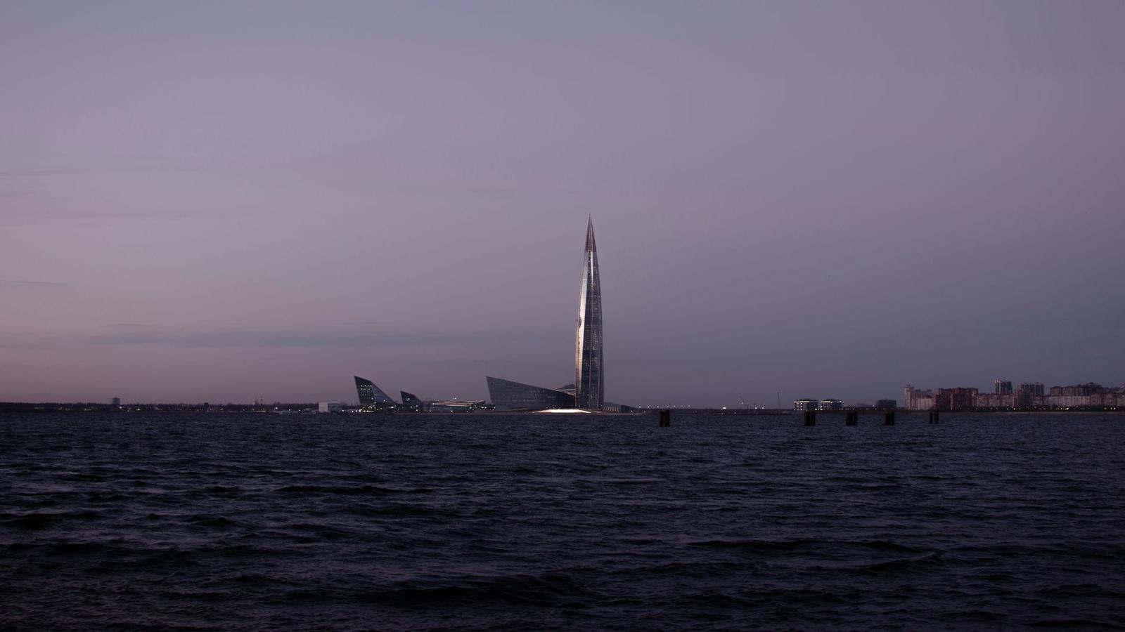 Slender glass skyscraper spire rising above a dusky waterfront