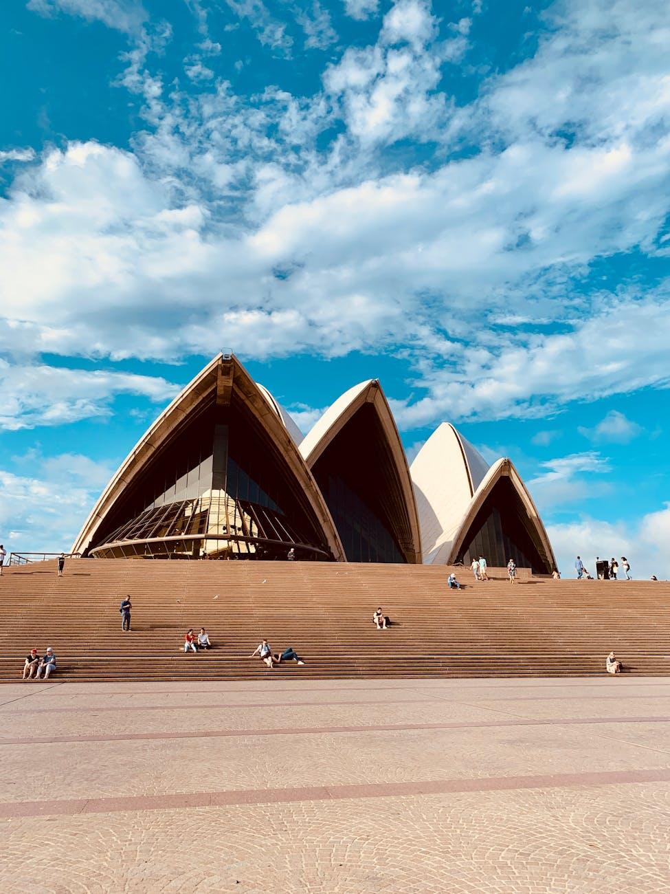 Traveling people walking around Opera House with triangle shaped roof on background of blue cloudy sky in summer in Sydney