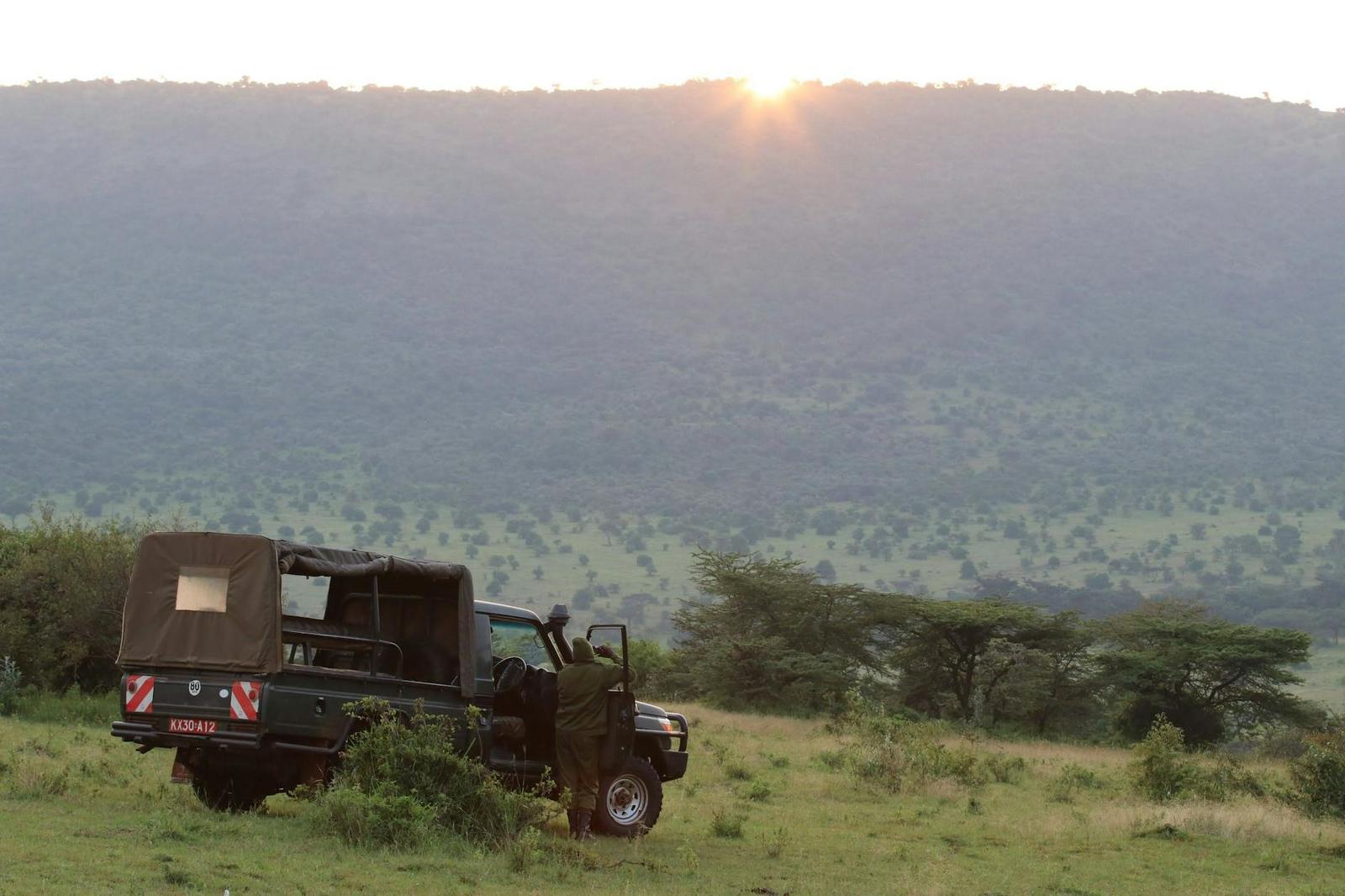 A safari vehicle parked on a grassy ridge as the sun rises over hills