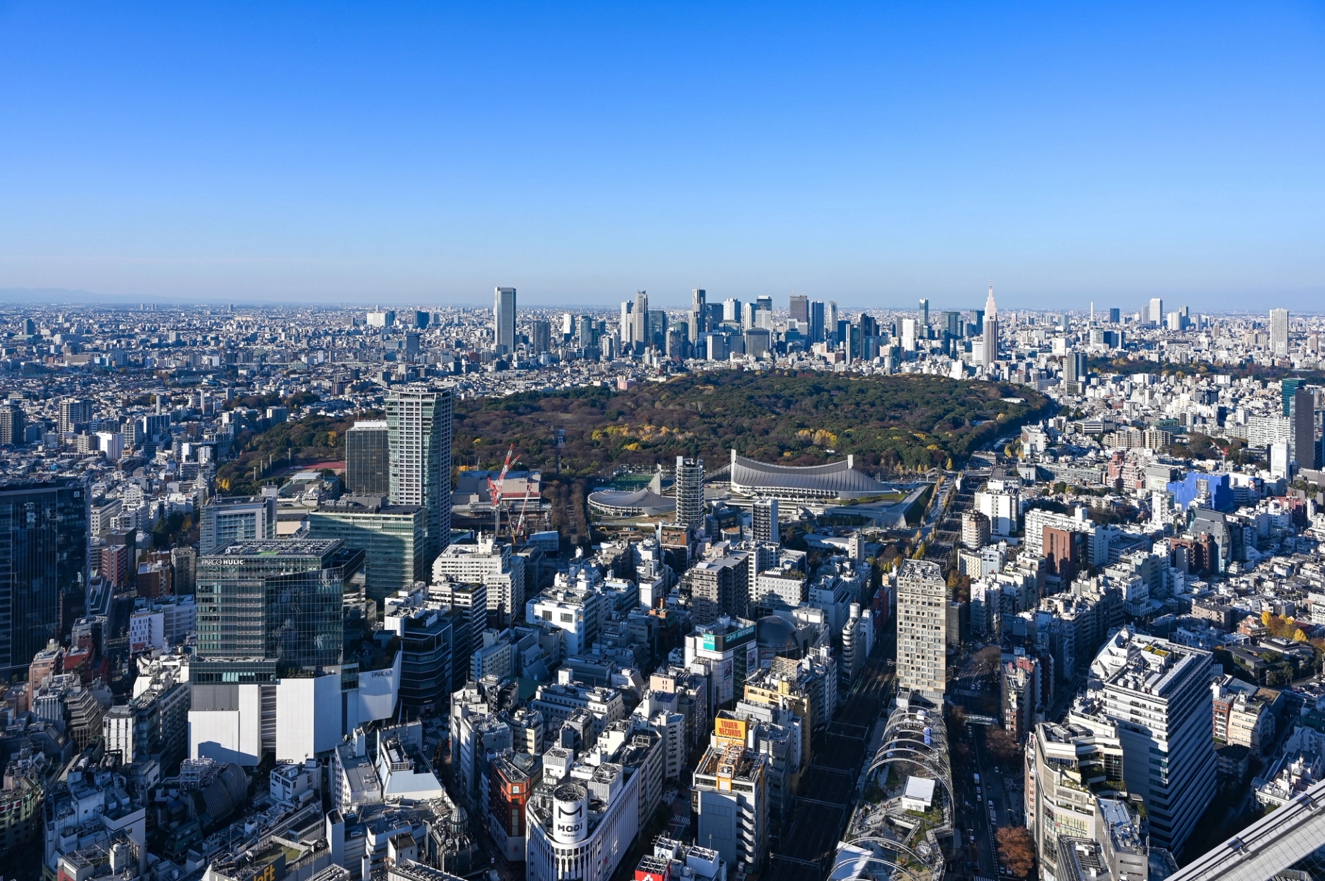 Aerial view of Tokyo cityscape featuring Shinjuku Park surrounded by skyscrapers under a clear blue sky