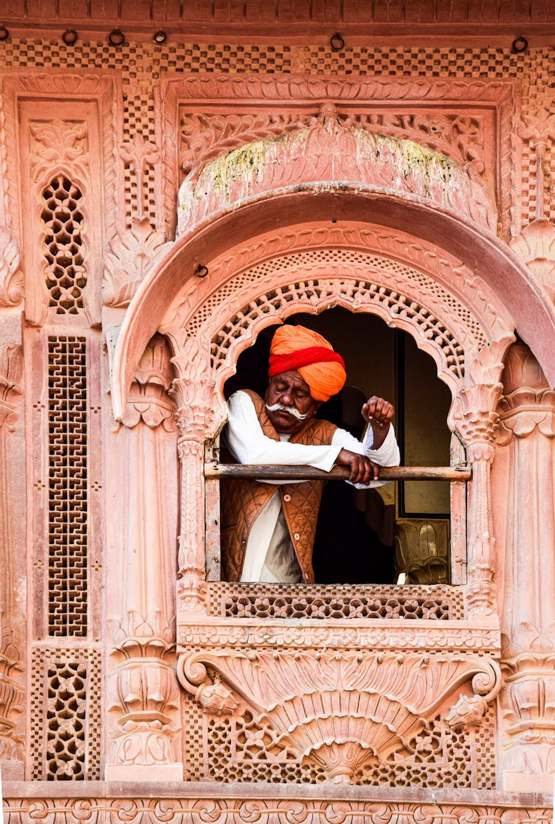 A senior Indian man in a turban leans from an ornate window of a traditional building