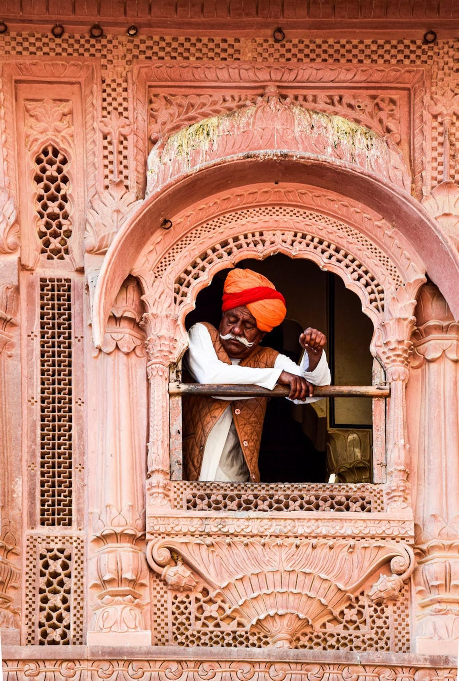A senior Indian man in a turban leans from an ornate window of a traditional building