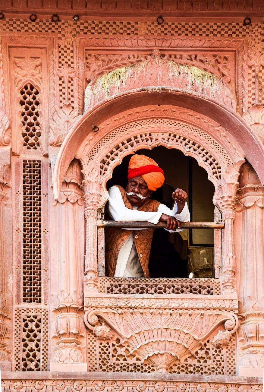 A senior Indian man in a turban leans from an ornate window of a traditional building