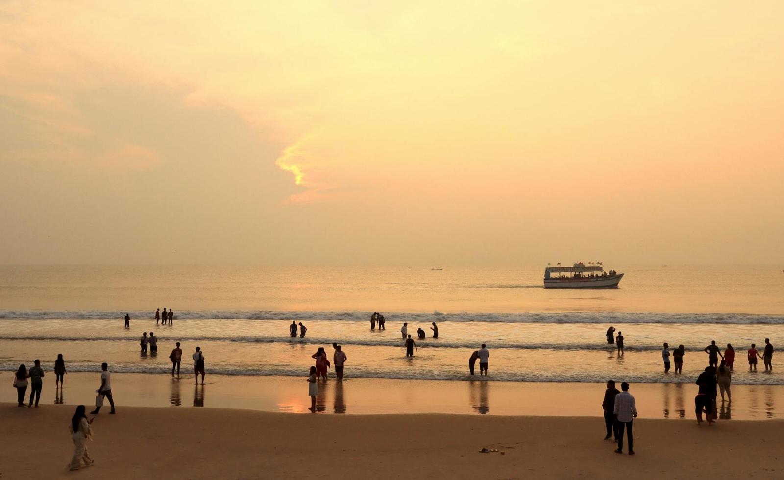 Silhouetted beachgoers wading in the surf as a boat passes at sunset