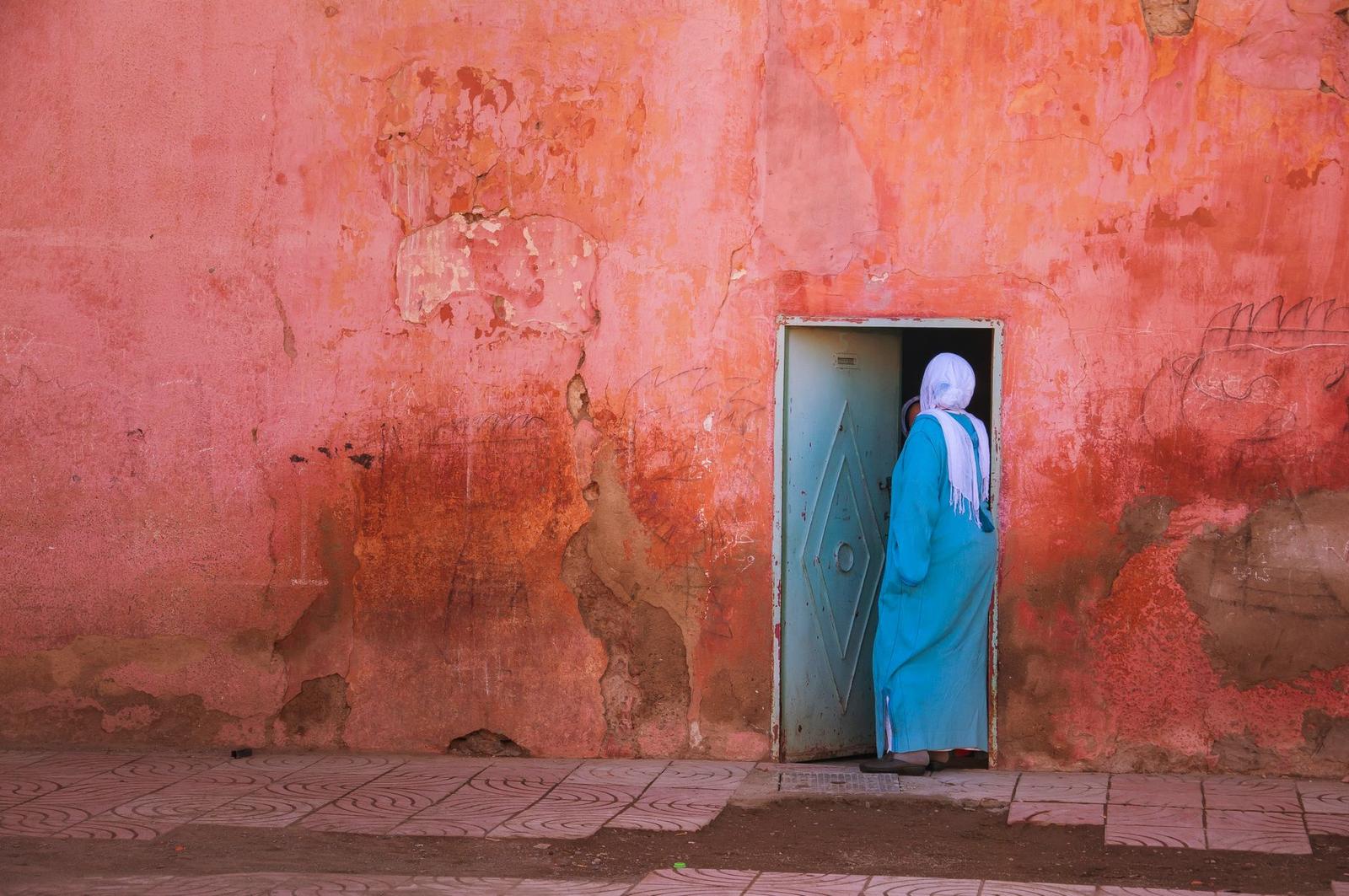 A woman in a headscarf and blue robe stepping through a teal door in a weathered pink wall