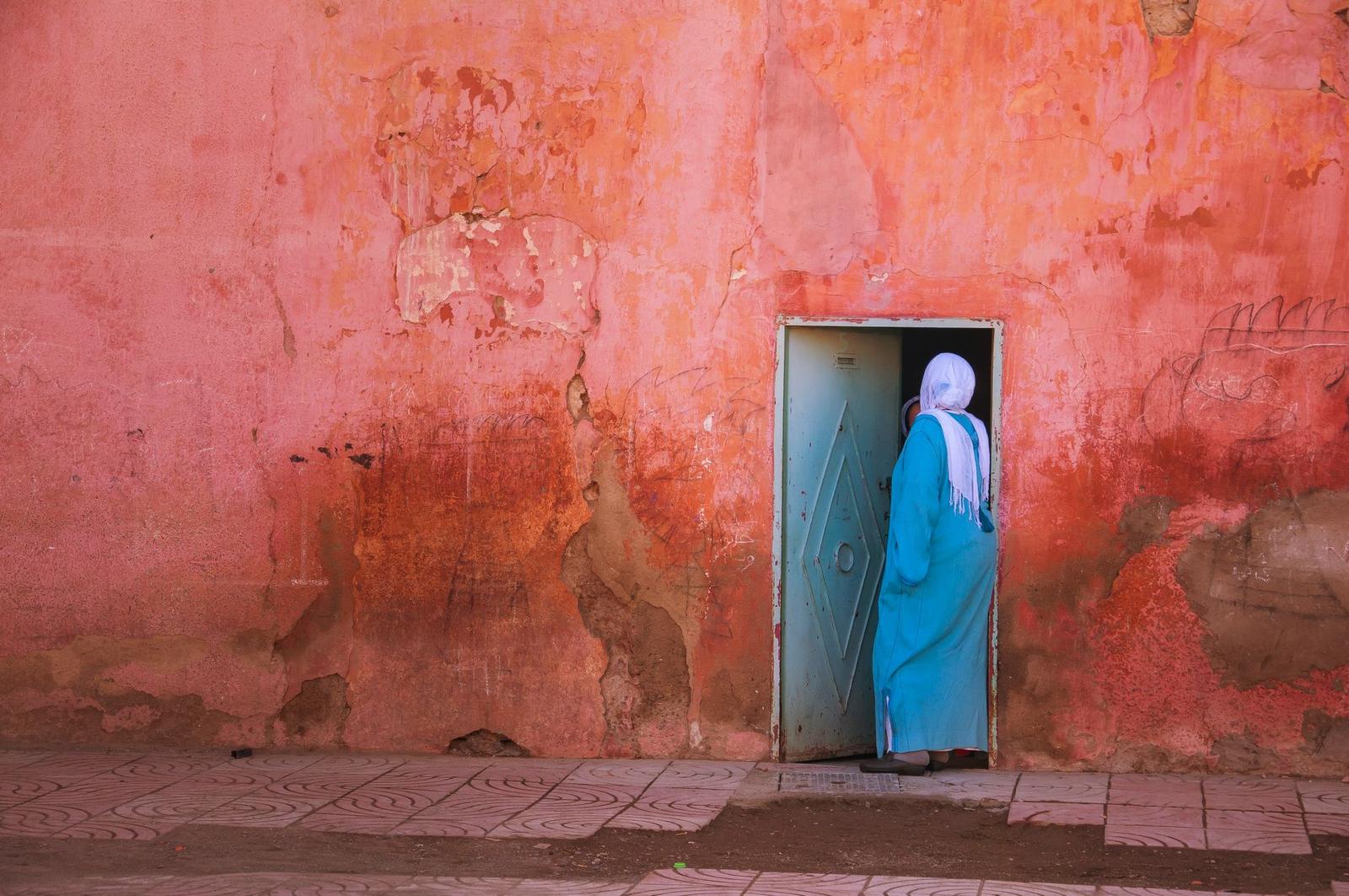 A woman in a headscarf and blue robe stepping through a teal door in a weathered pink wall