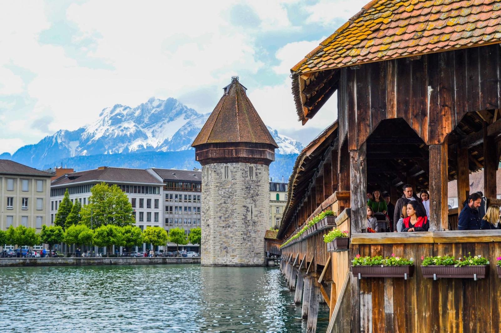 Group of tourists walking on wooden bridge over calm river located near old brick tower against mountains in daytime