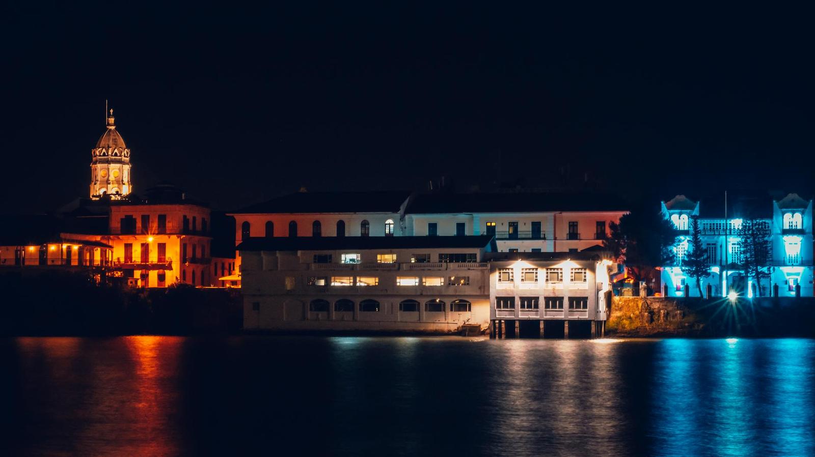 A lit waterfront building with a corner tower glowing against a dark lagoon at night