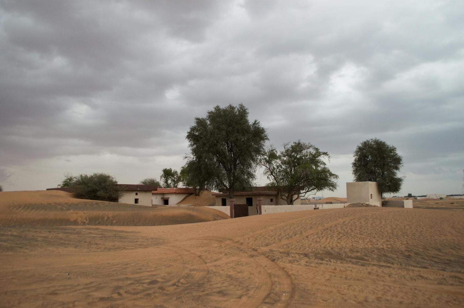 A small desert compound with trees and low buildings under a heavy grey sky