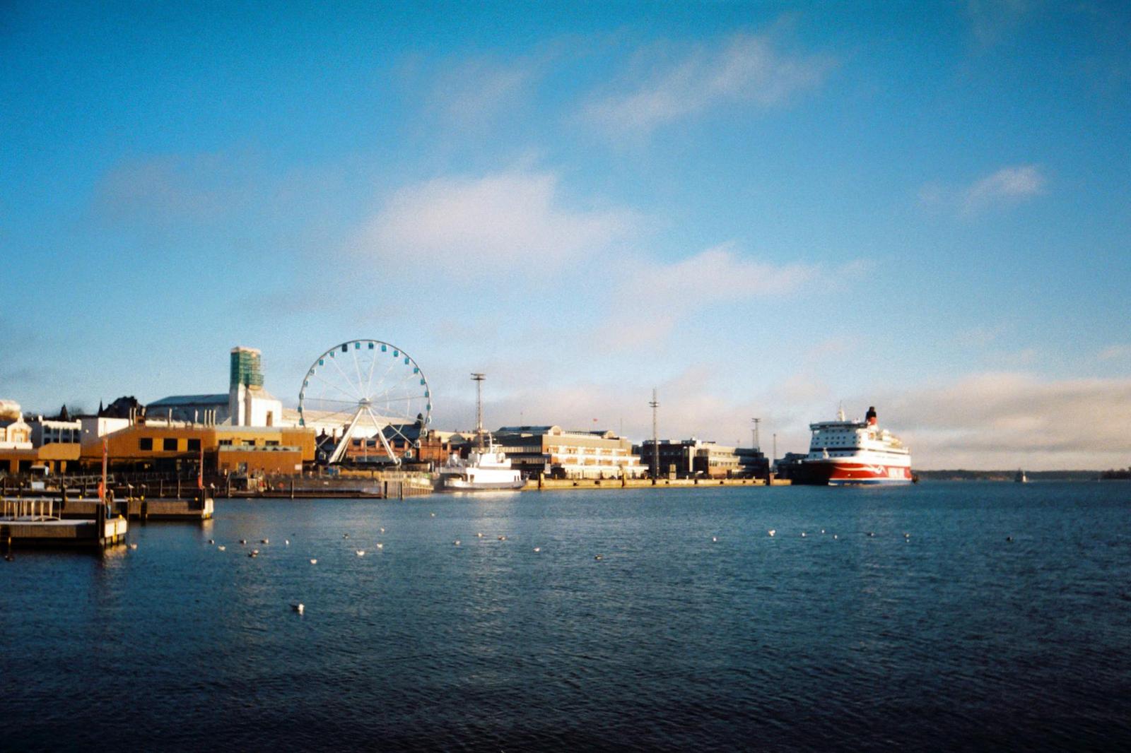 A waterfront skyline with a ferris wheel and cruise liner under a pale blue sky
