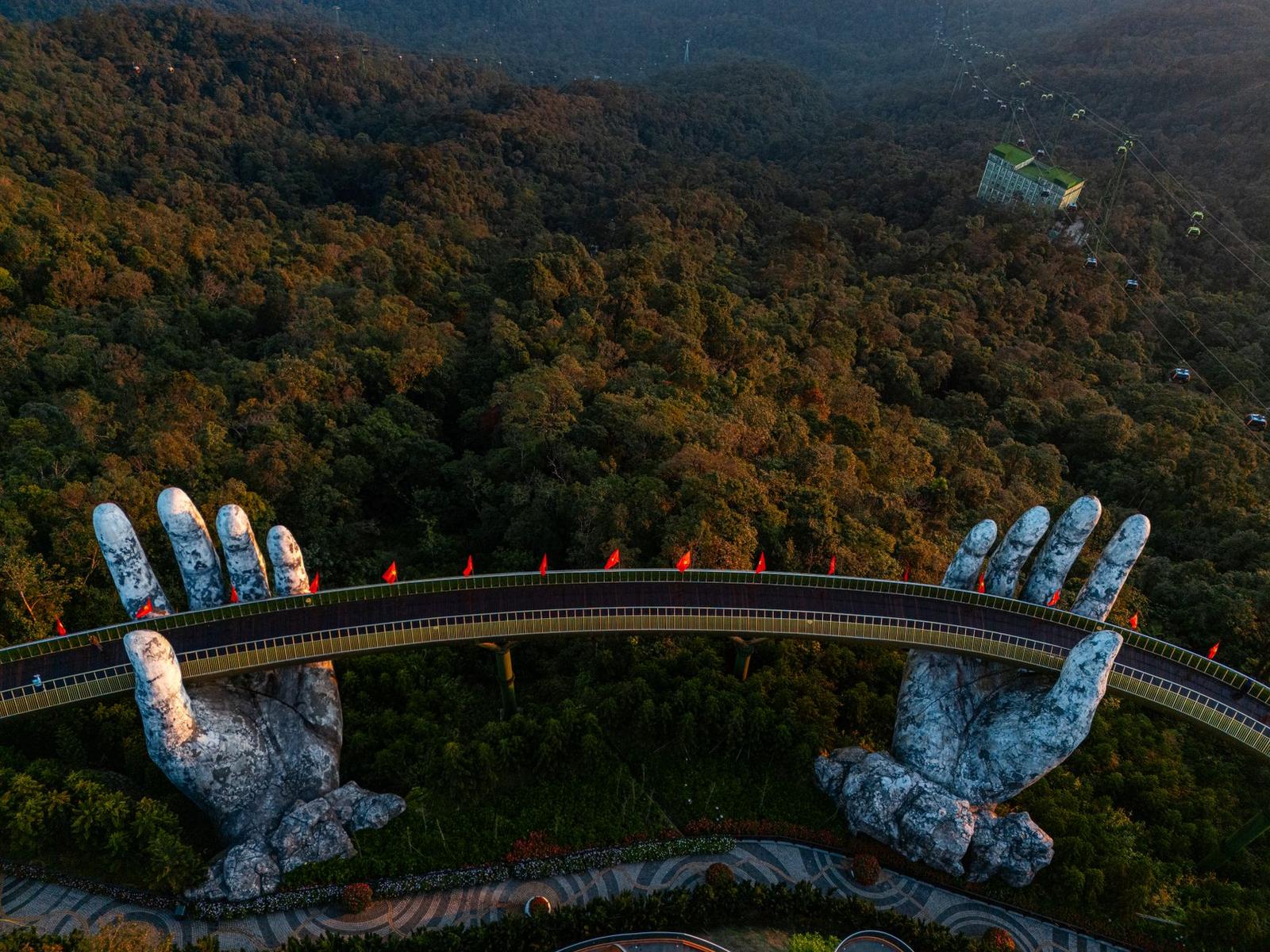 Giant stone hand sculptures cradling a golden footbridge over a forested mountain