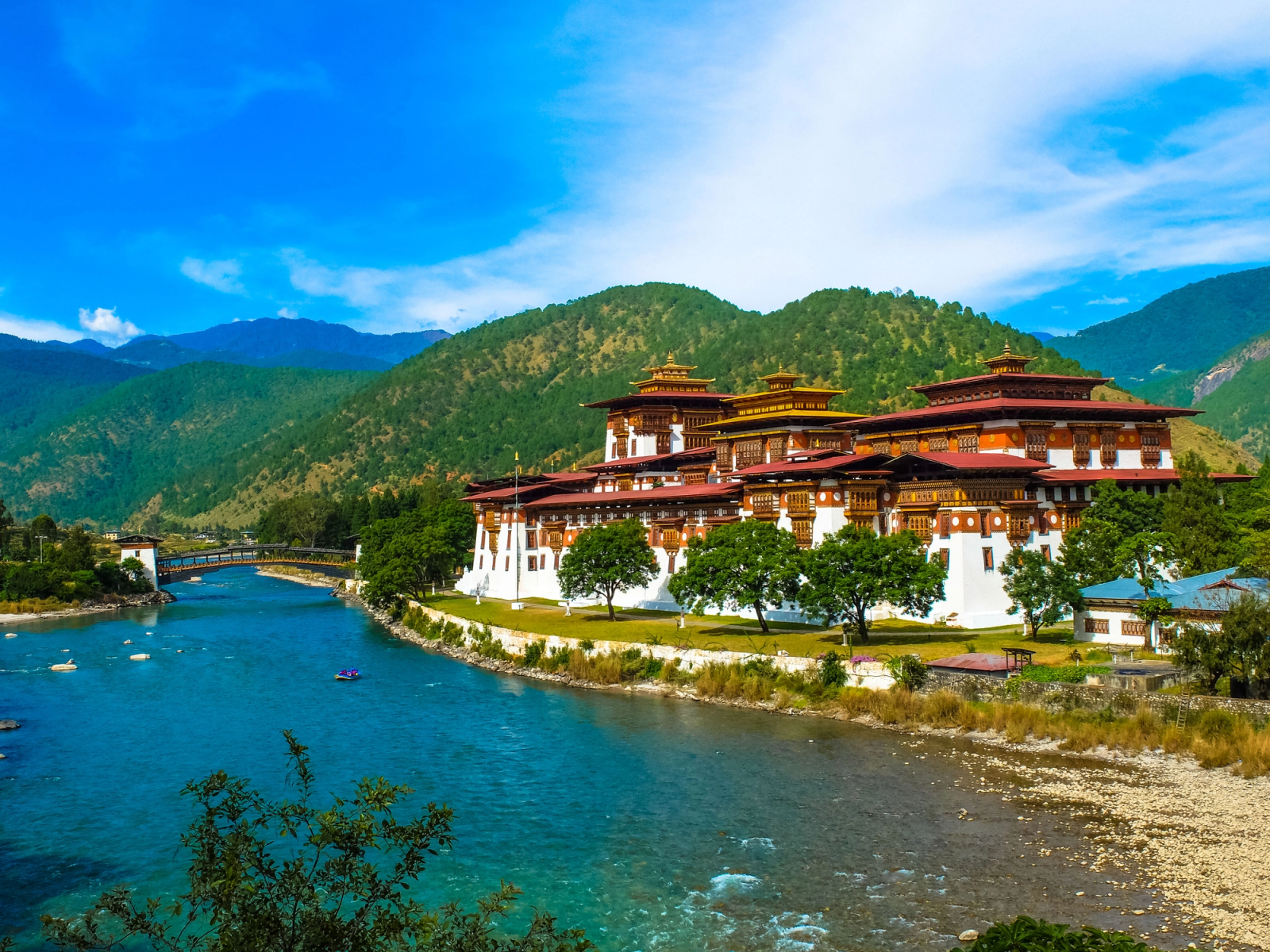 A grand white fortress-monastery beside a river with a wooden bridge and forested hills