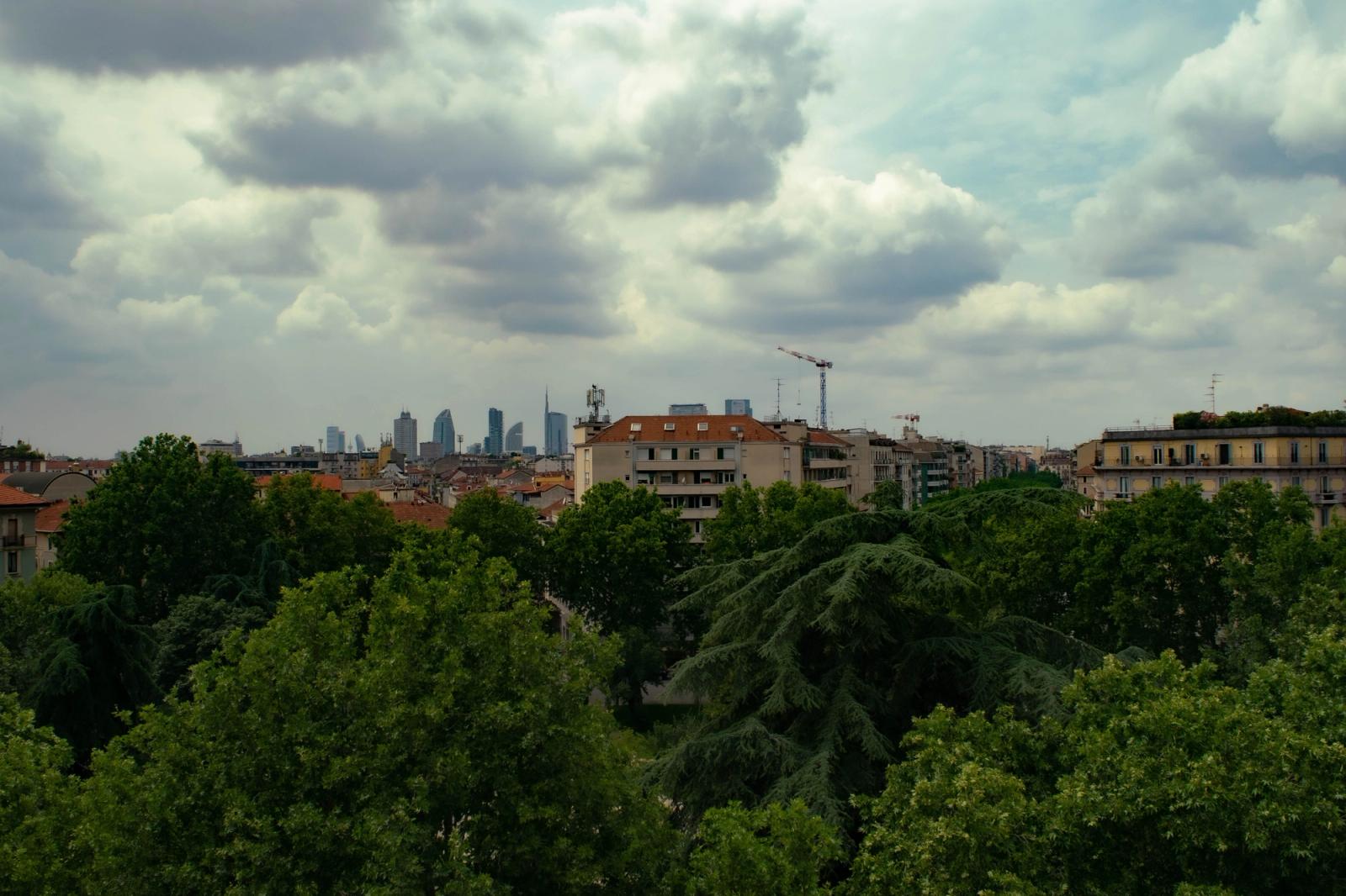 Skyline of Milan with clear view of urban buildings amidst lush green trees under a cloudy sky