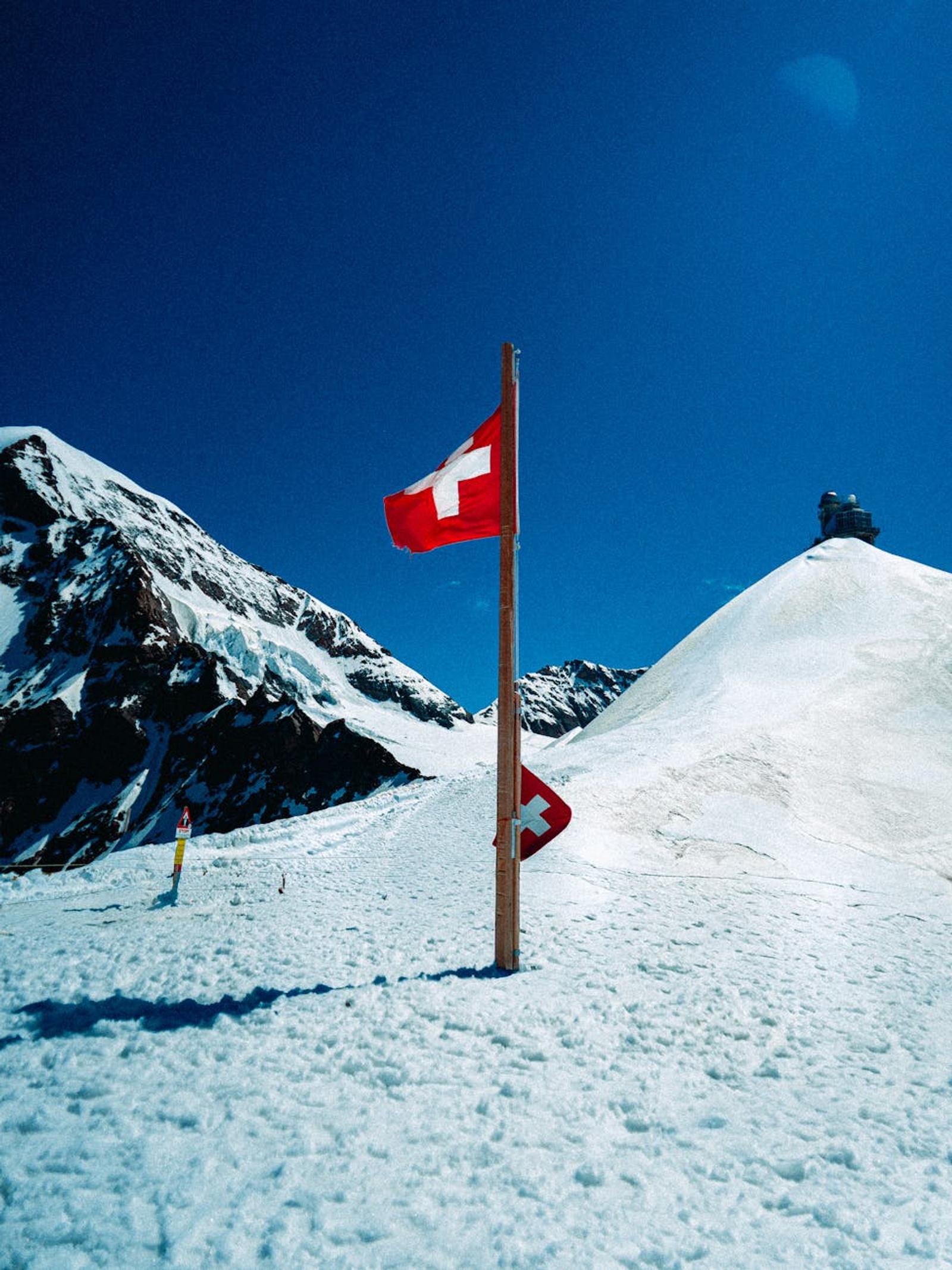 A Swiss flag on a snowy mountain peak with a clear blue sky