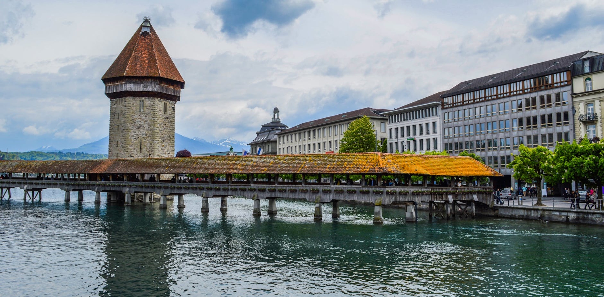 Stone water tower and covered wooden bridge spanning a river beside a row of townhouses