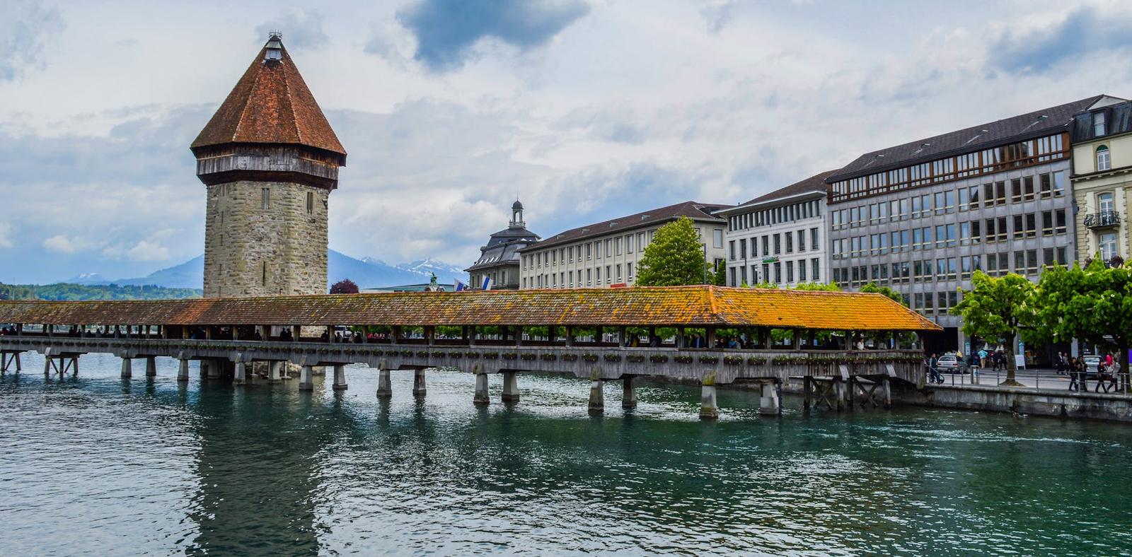 Stone water tower and covered wooden bridge spanning a river beside a row of townhouses