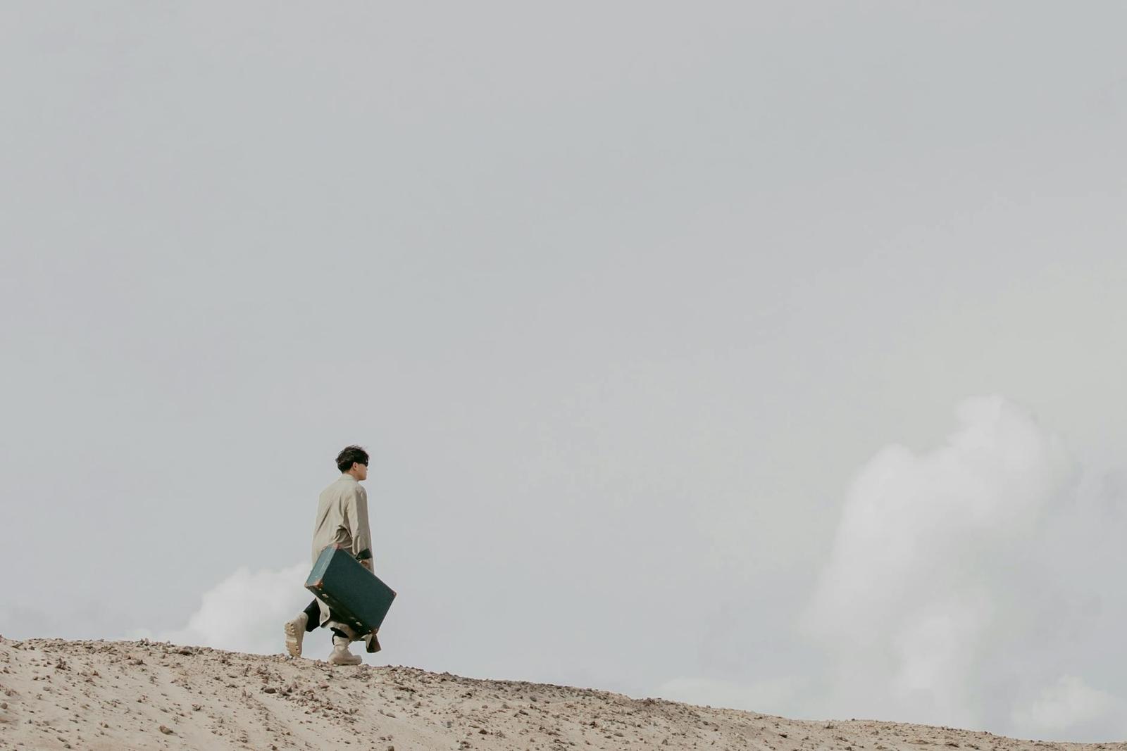 A distant figure walks across a sandy dune carrying a suitcase