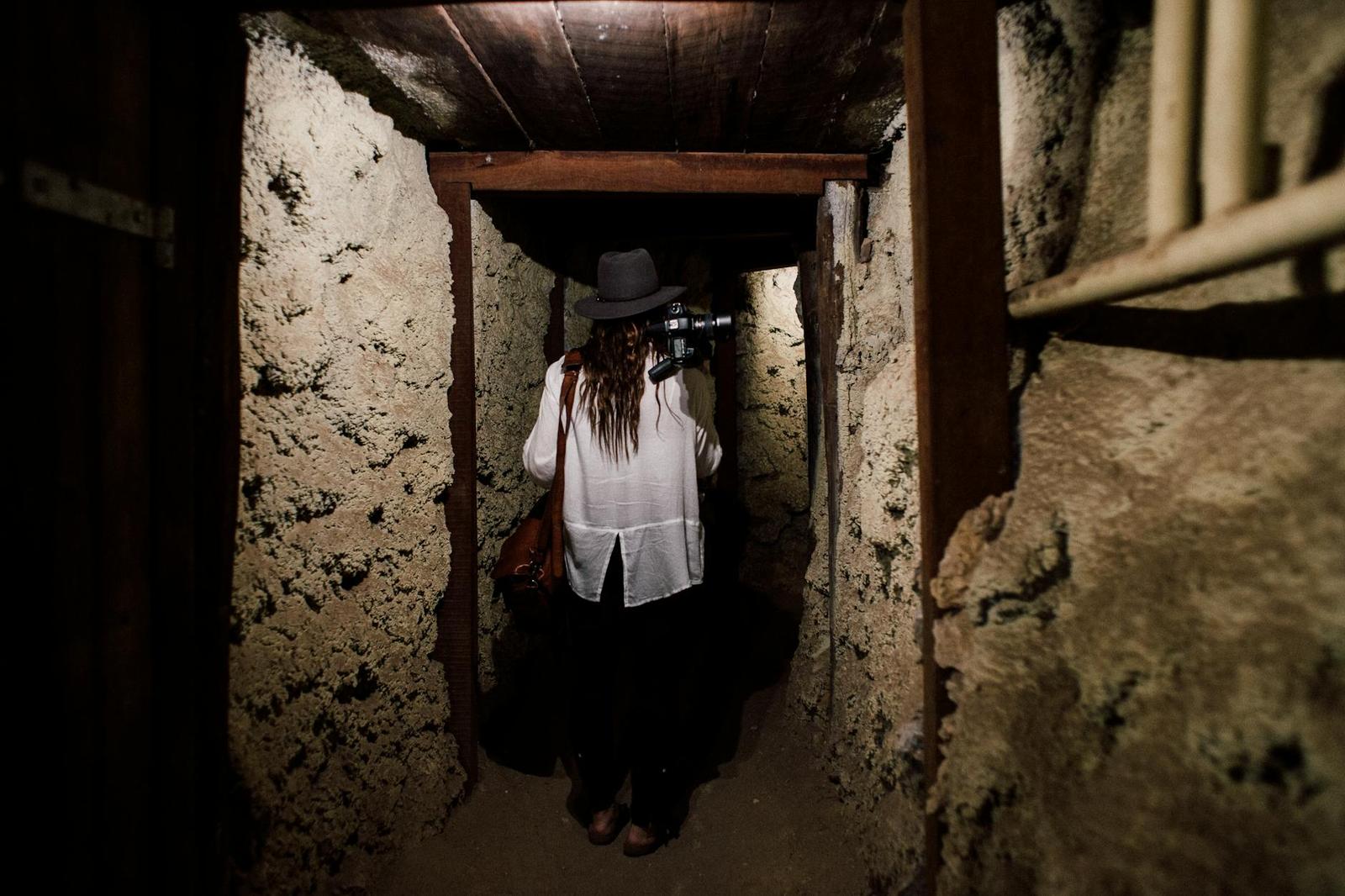 A woman in a white blouse and hat walking through a narrow dug-earth tunnel