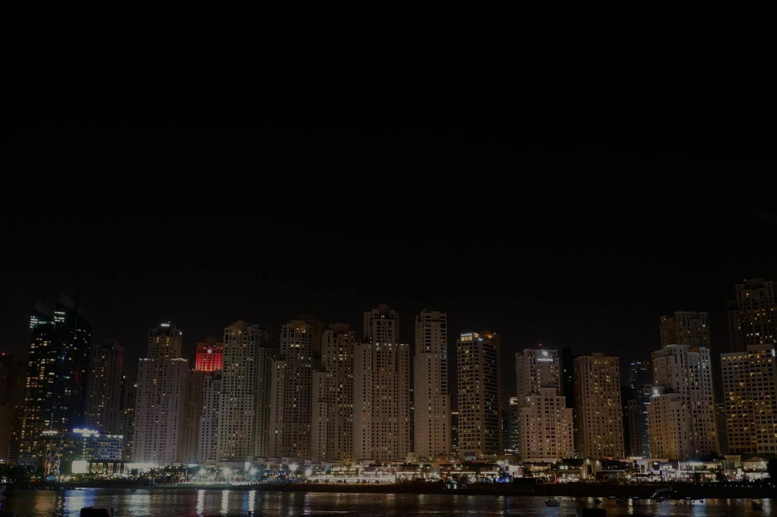 A dark city skyline of lit towers along the waterfront at night