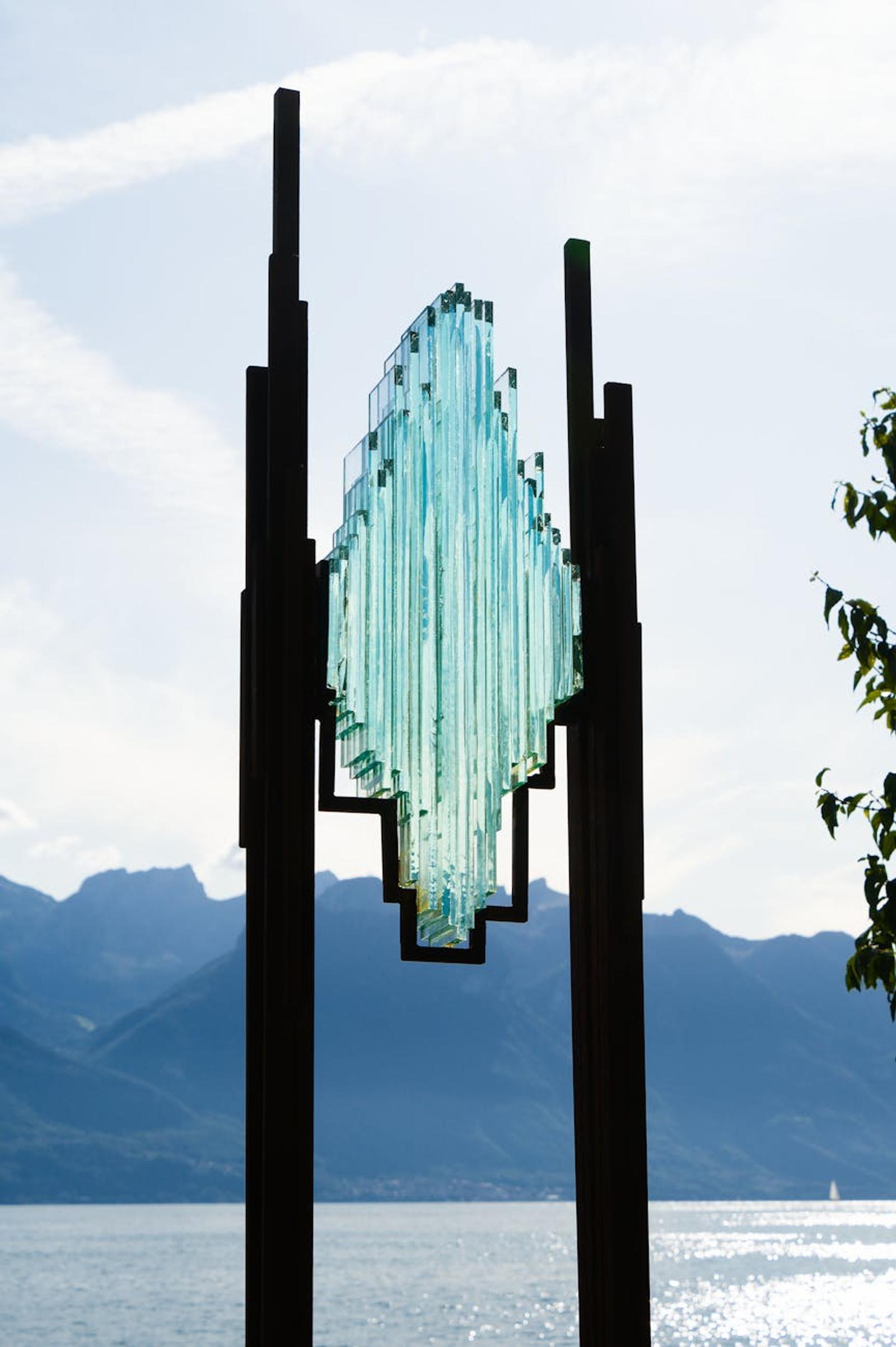 Glass sculpture on metal posts silhouetted against a lakeside mountain view