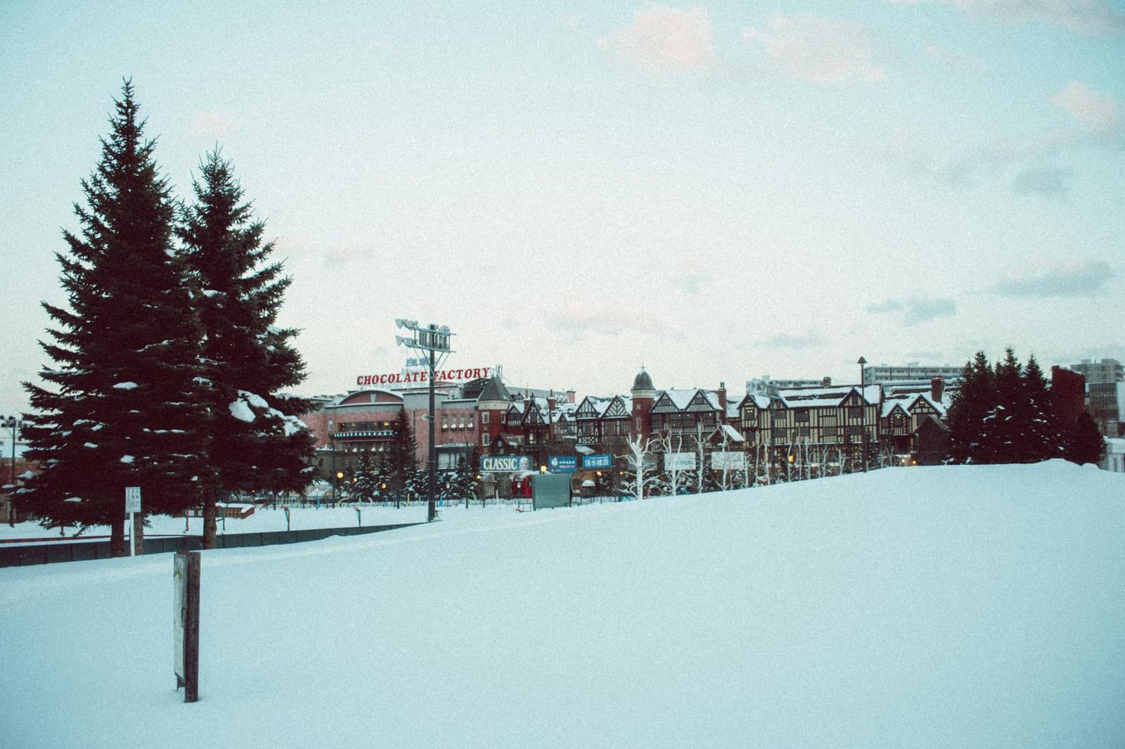 Snow-covered plaza with pine trees and a row of alpine buildings under a pale sky