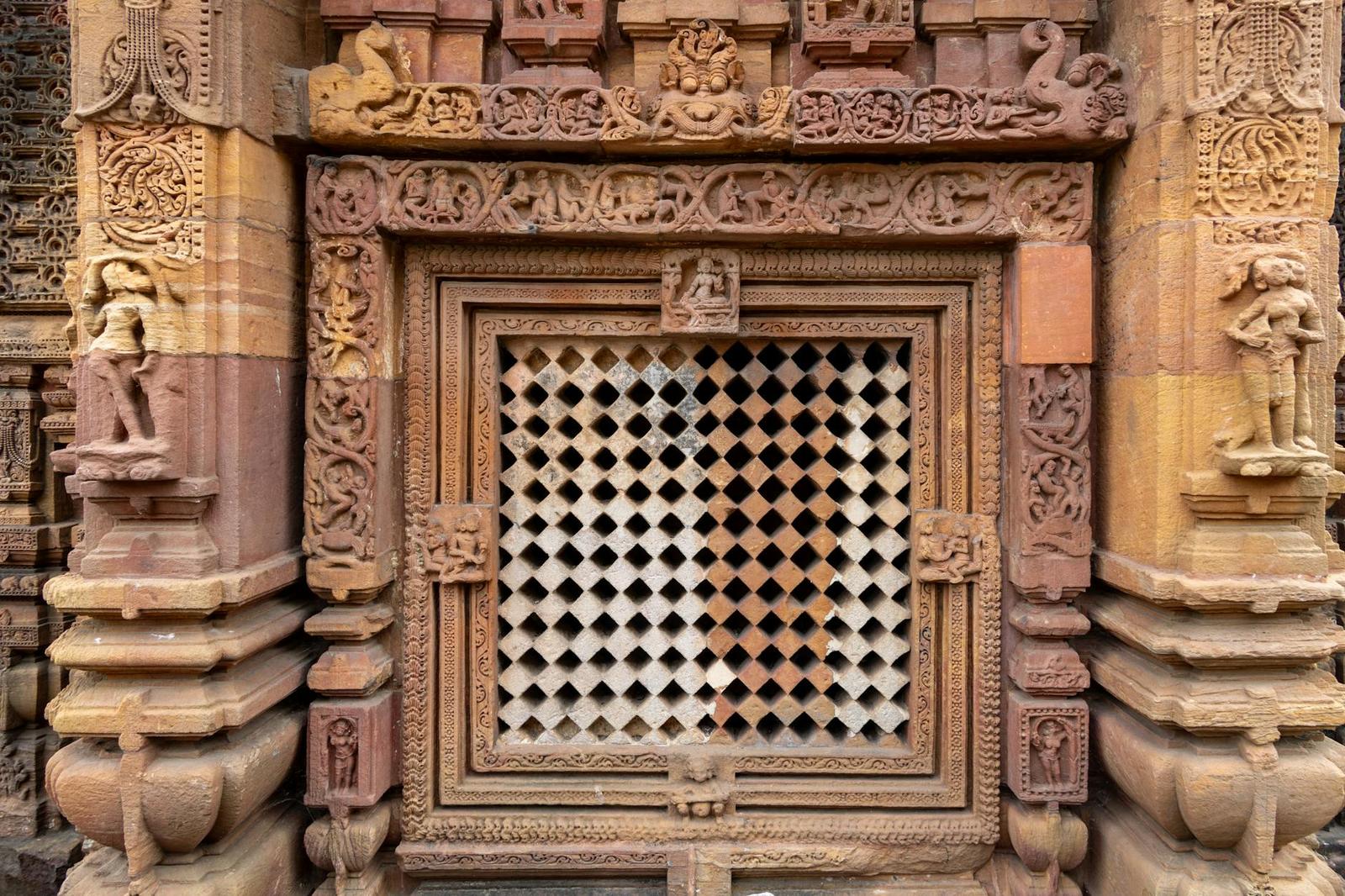 A carved stone temple window with a diamond lattice screen flanked by sculpted figures