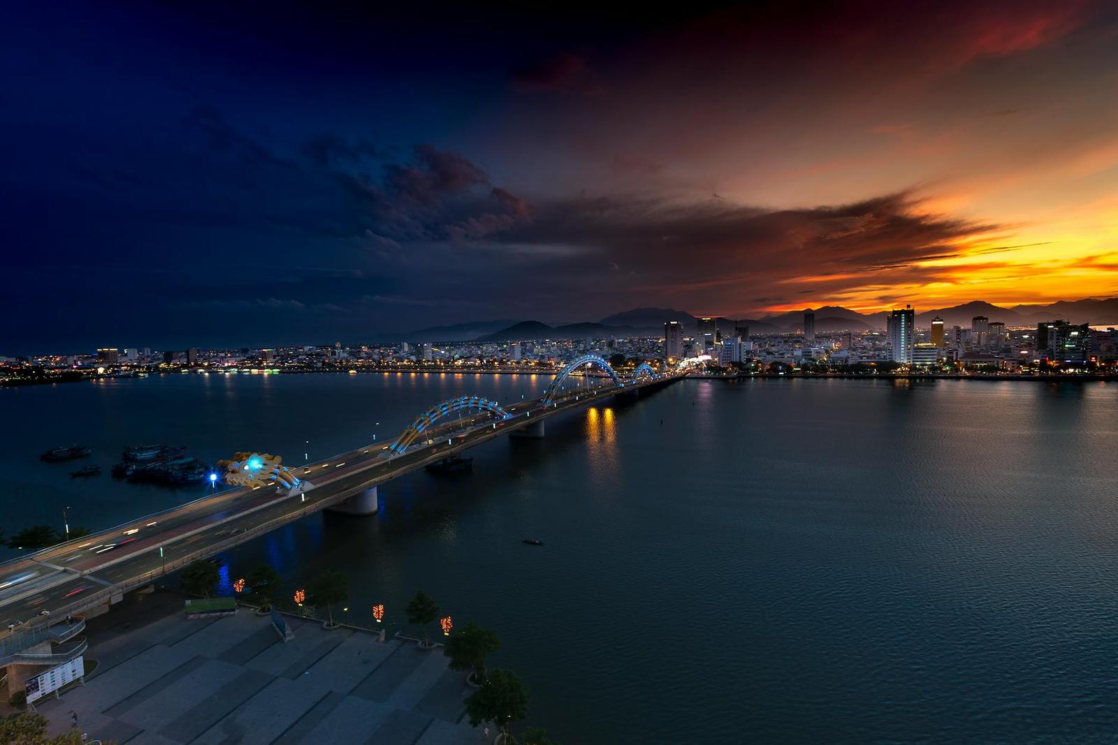 Illuminated bridge crossing a river beneath a dramatic dusk sky