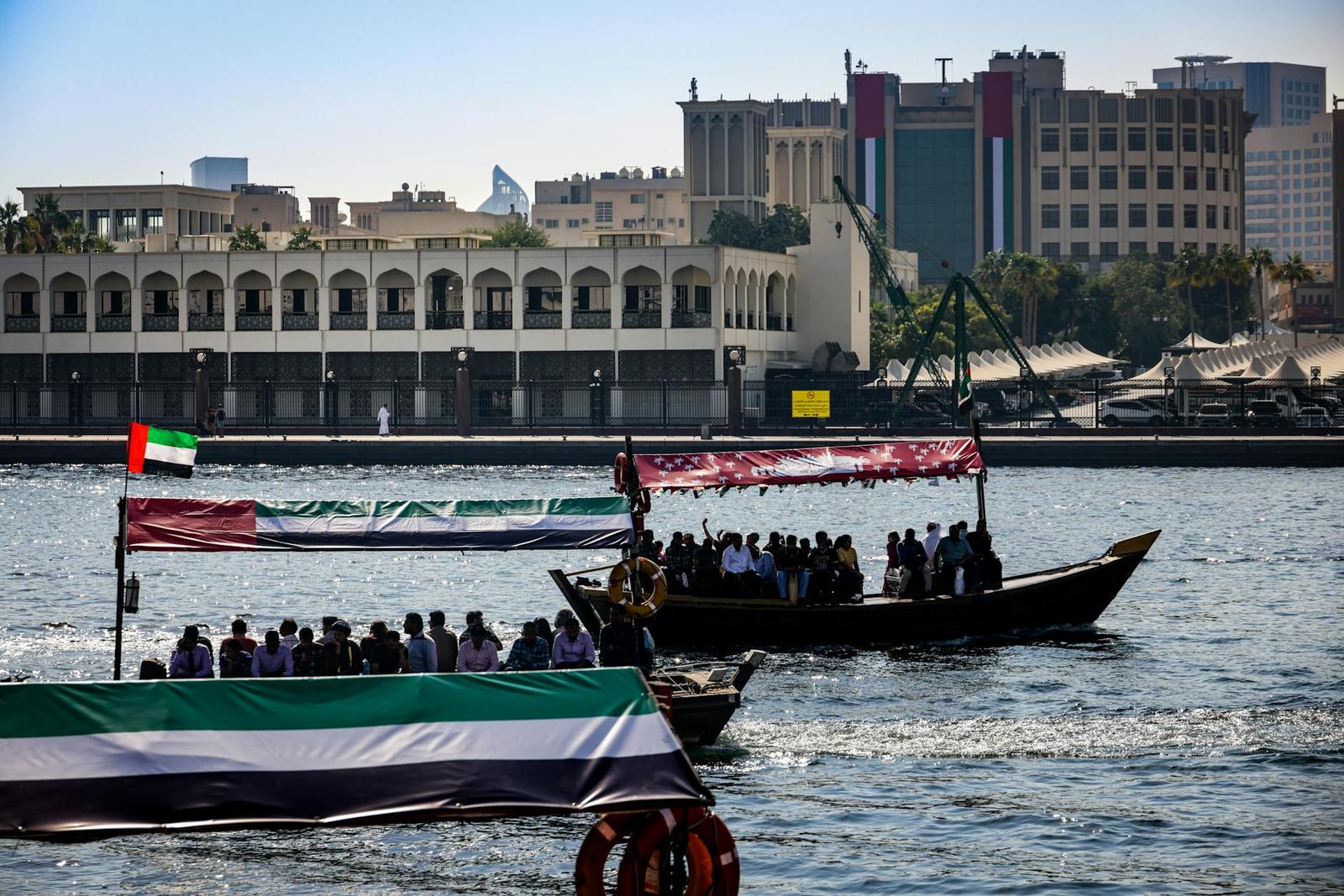 Passenger boats decked with flags crossing a creek past heritage buildings