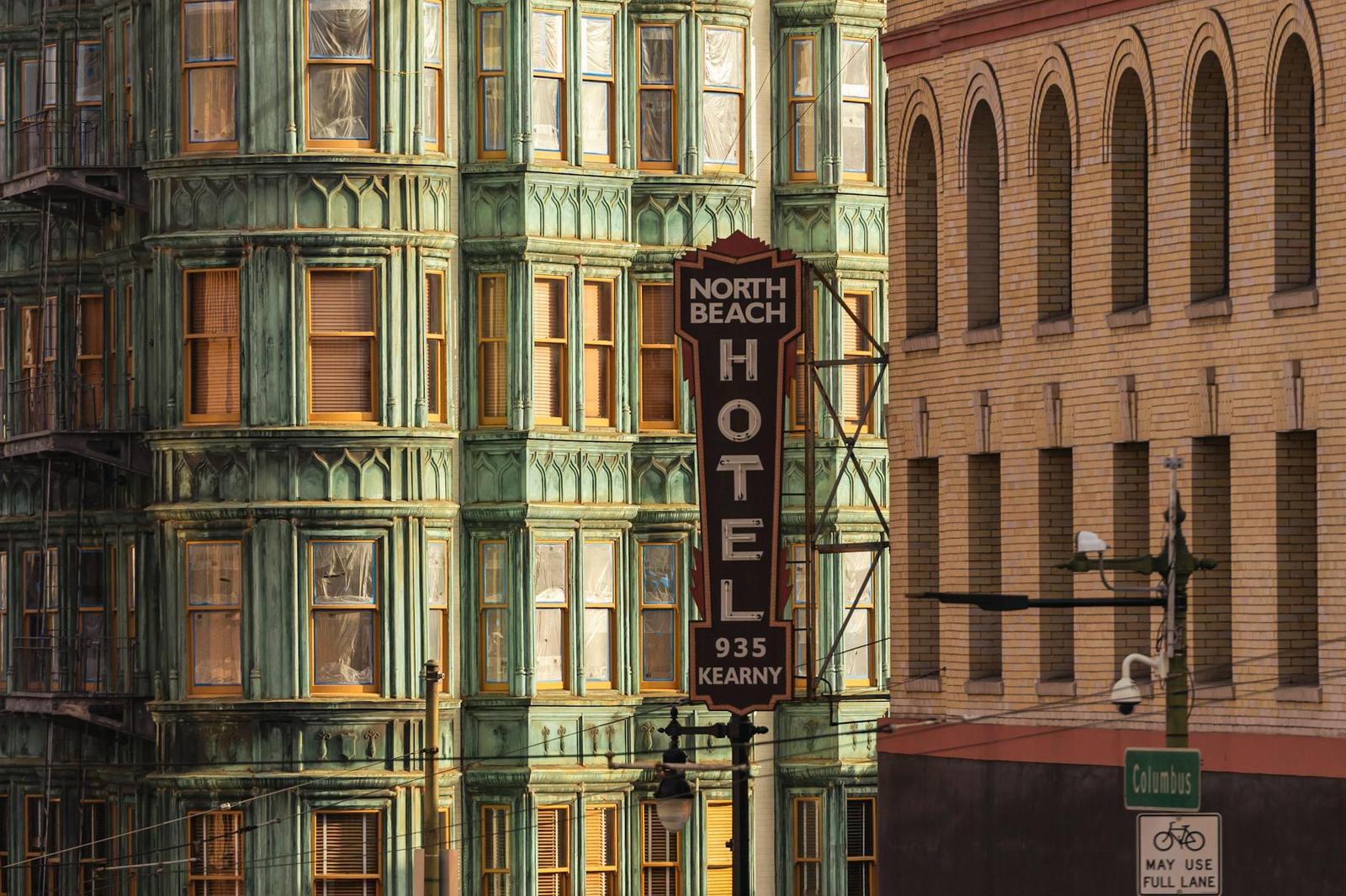 A green bay-windowed hotel building with a vertical hotel sign beside a brick facade