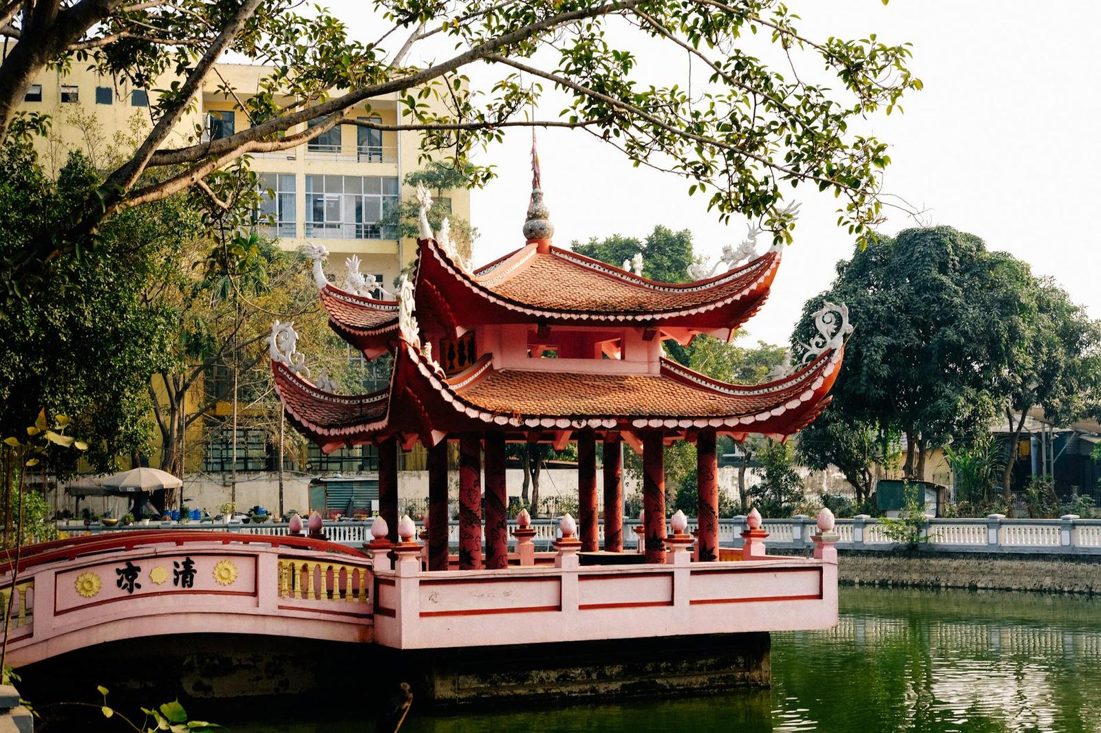 Red-roofed lakeside pavilion connected by a curved bridge among city trees
