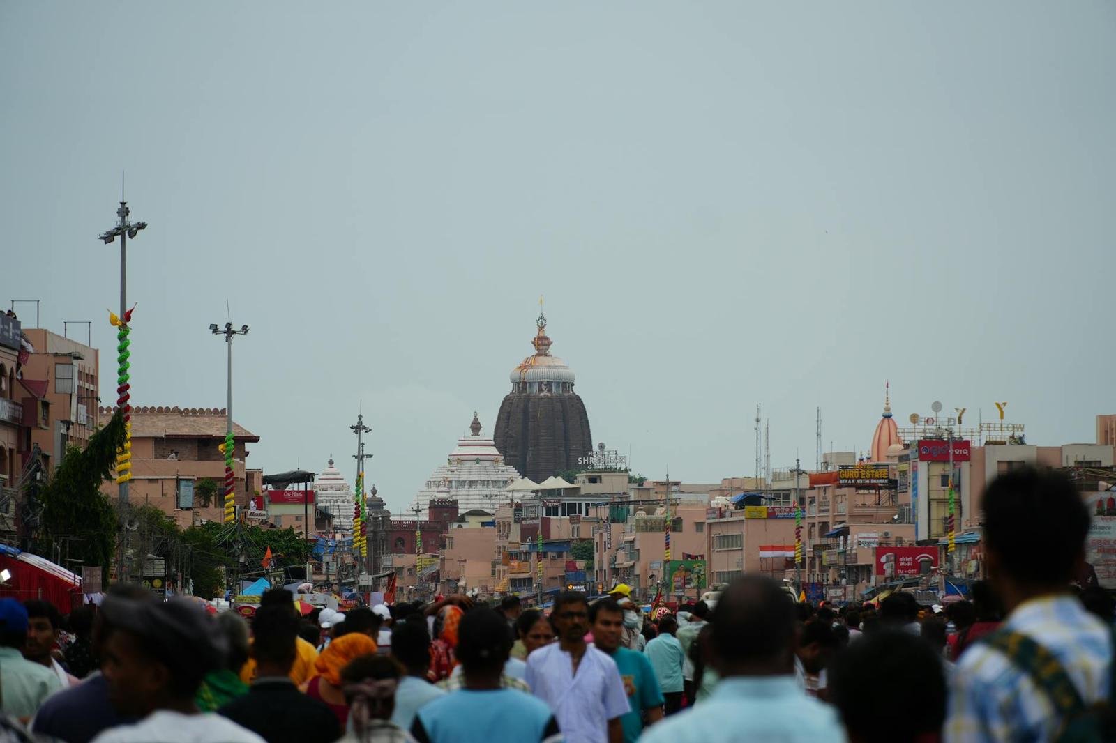 A crowd filling a street beneath a distant domed temple tower under grey skies