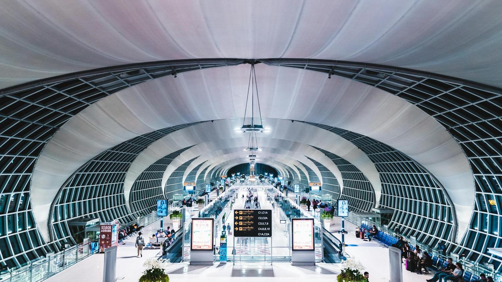 A curved glass and steel airport terminal with travelers below departure boards