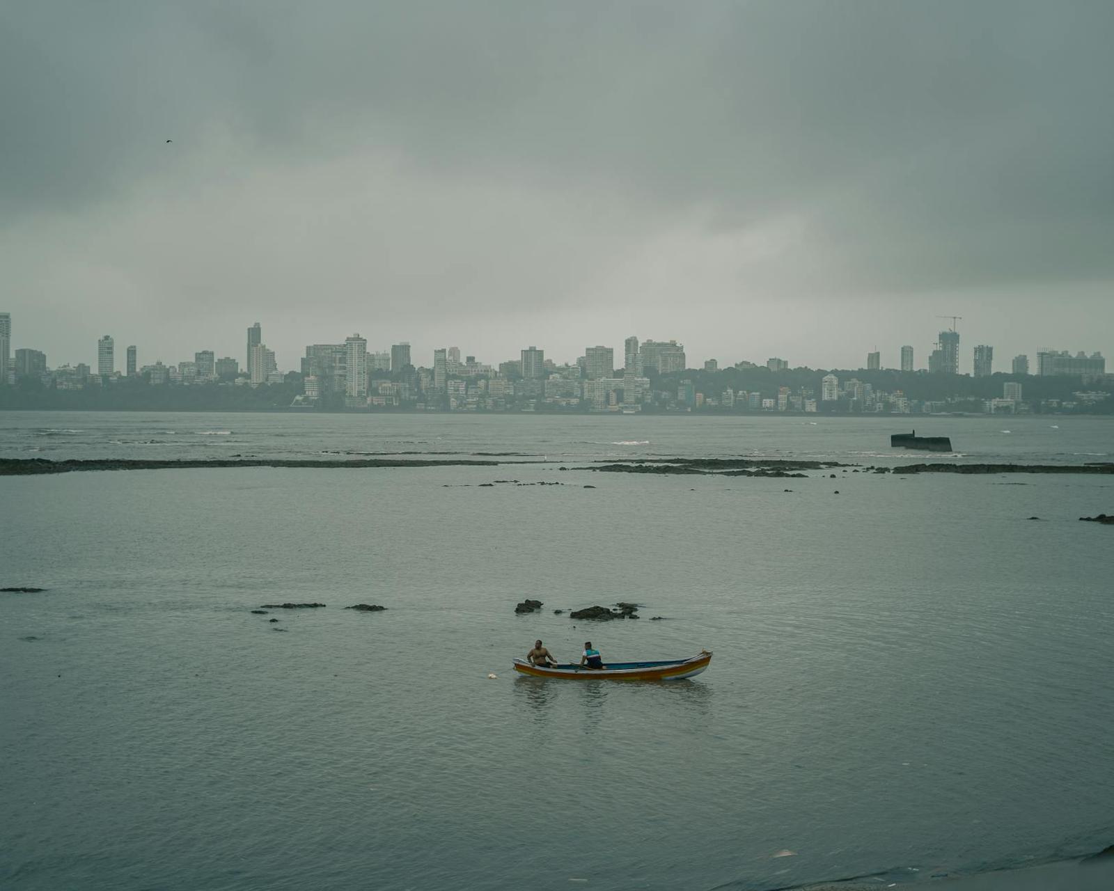 Two people in a small boat on calm water before a hazy city skyline