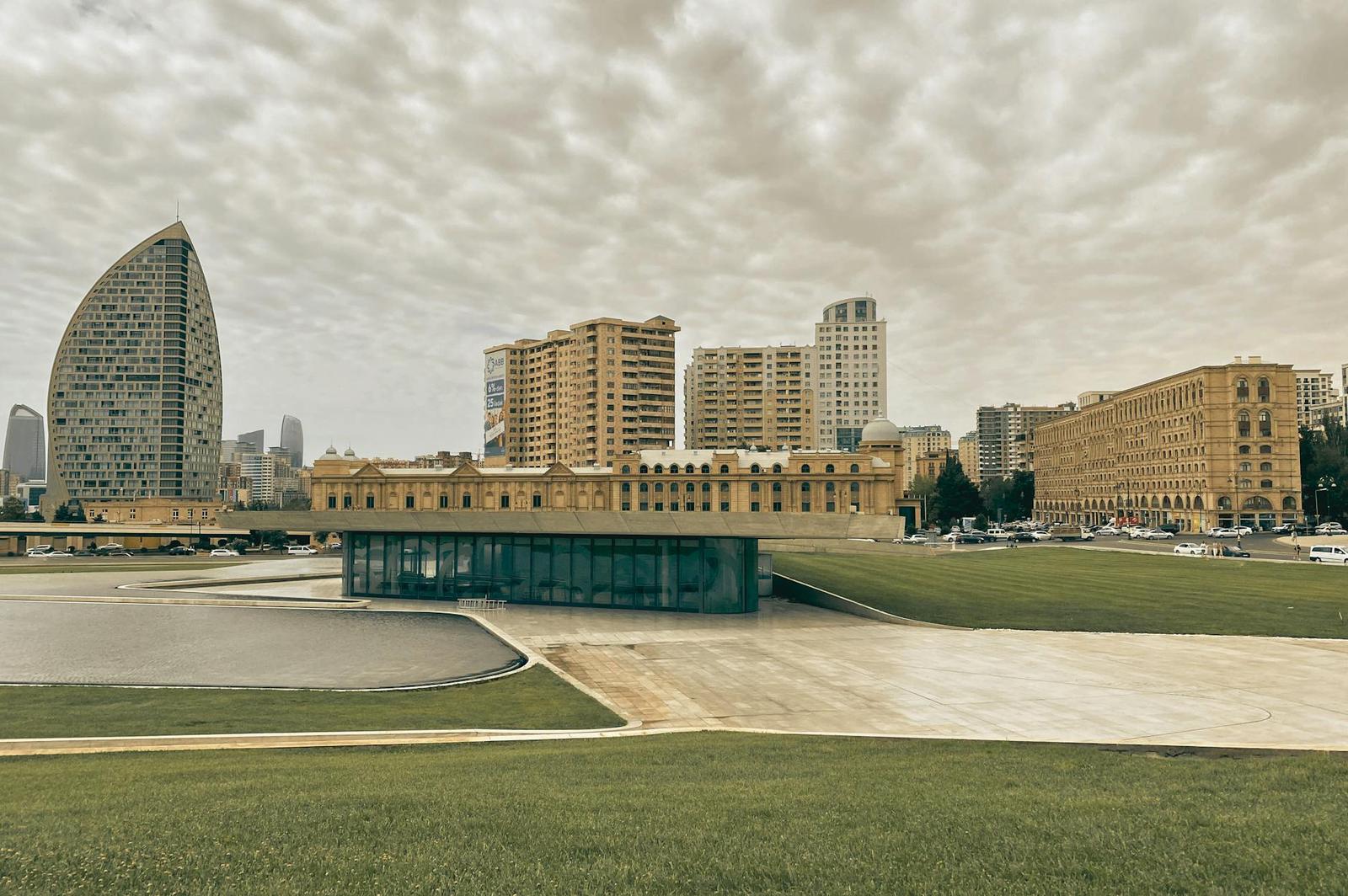 A glass pavilion sits on an open plaza beneath a sail-shaped skyscraper