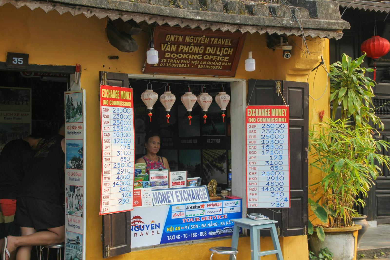A woman behind the counter of a yellow money exchange storefront hung with lanterns