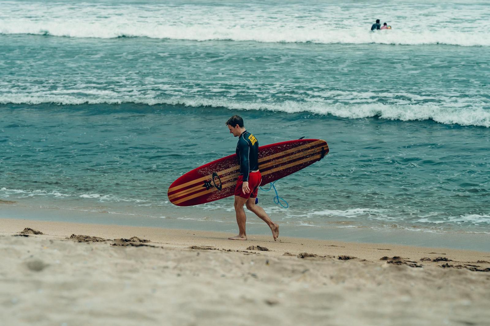 A surfer carries his board along the shoreline as waves roll in