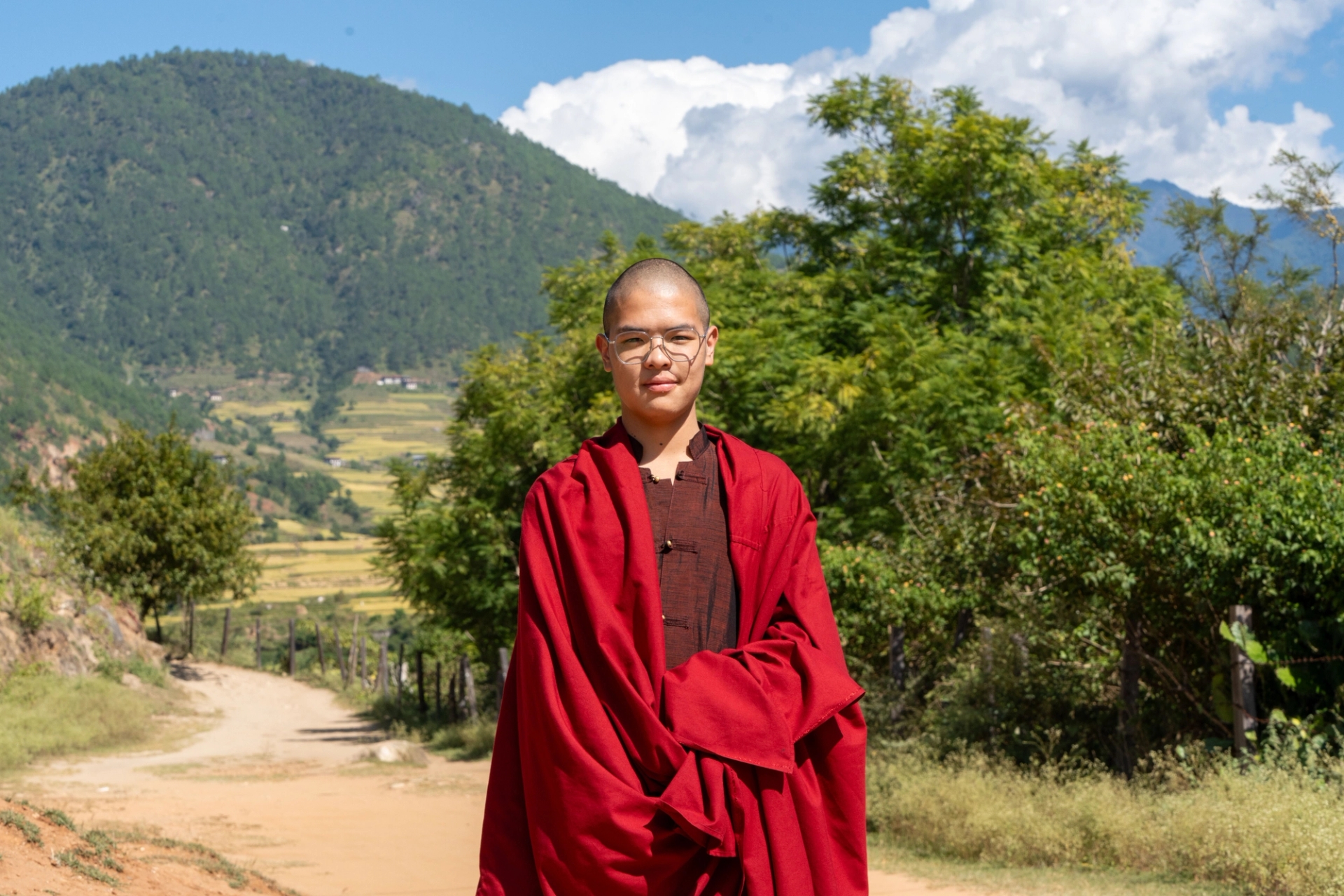 Monk in traditional red robes standing in a serene mountain landscape under a clear blue sky