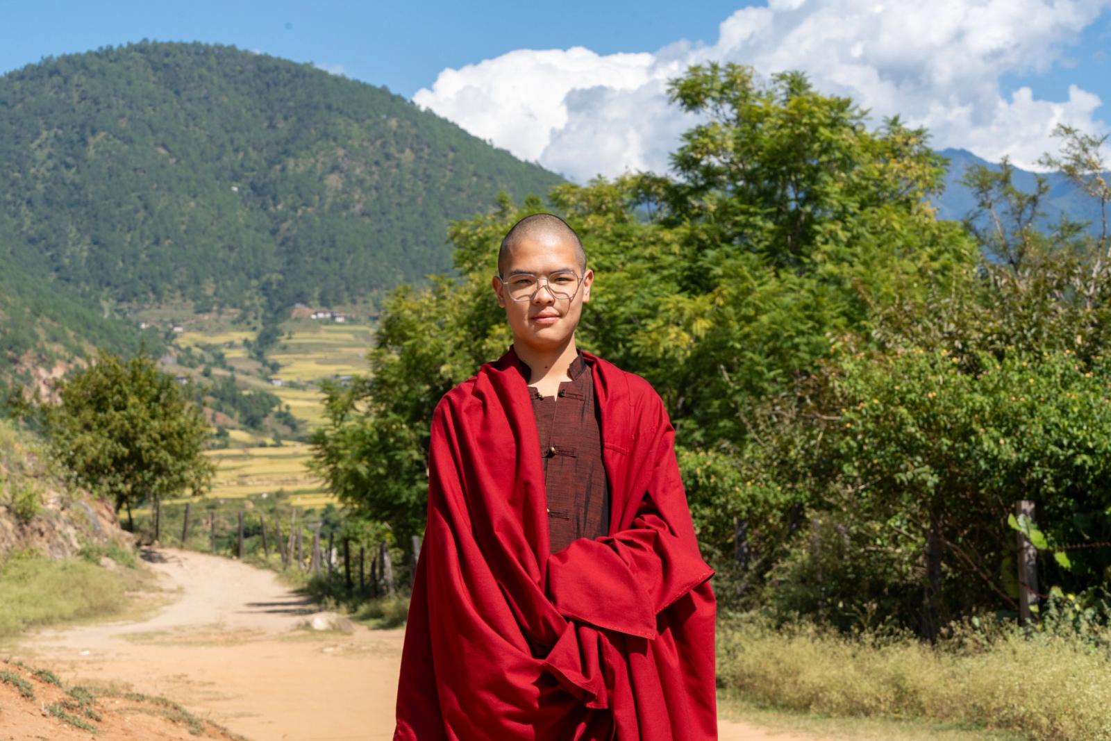 Monk in traditional red robes standing in a serene mountain landscape under a clear blue sky