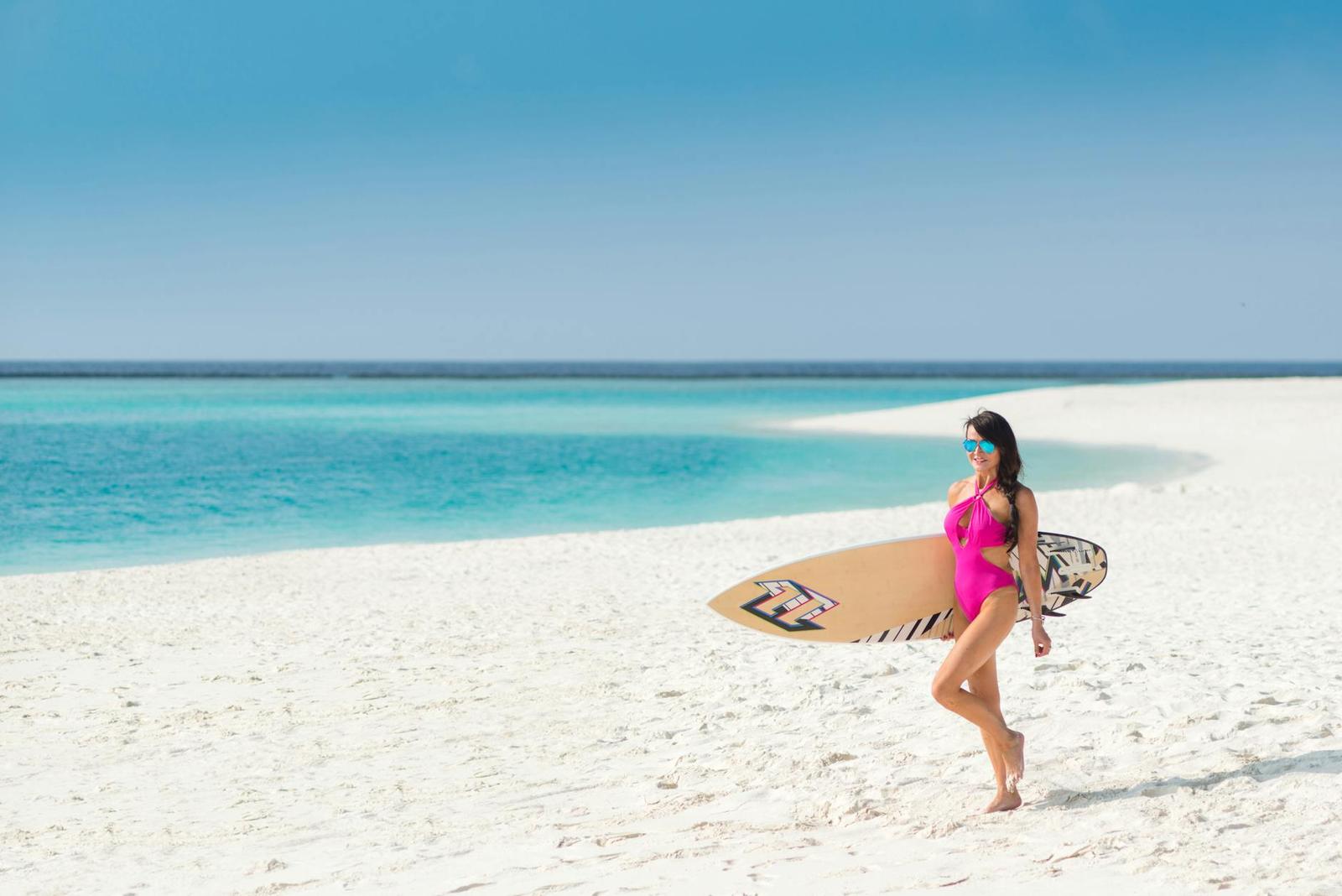 A woman in a pink swimsuit carrying a surfboard along a white sand beach