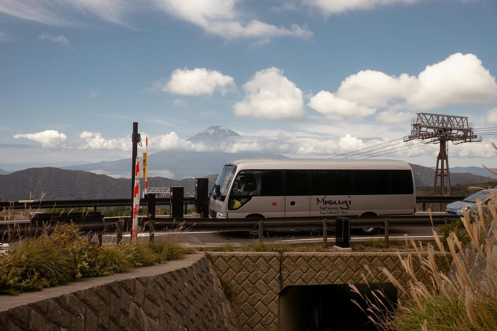 A shuttle bus on a mountain road beneath a cable car tower and distant peaks