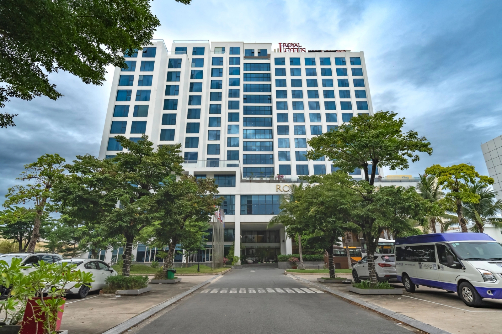 Front view of a modern hotel building surrounded by trees and parked vehicles under a cloudy sky