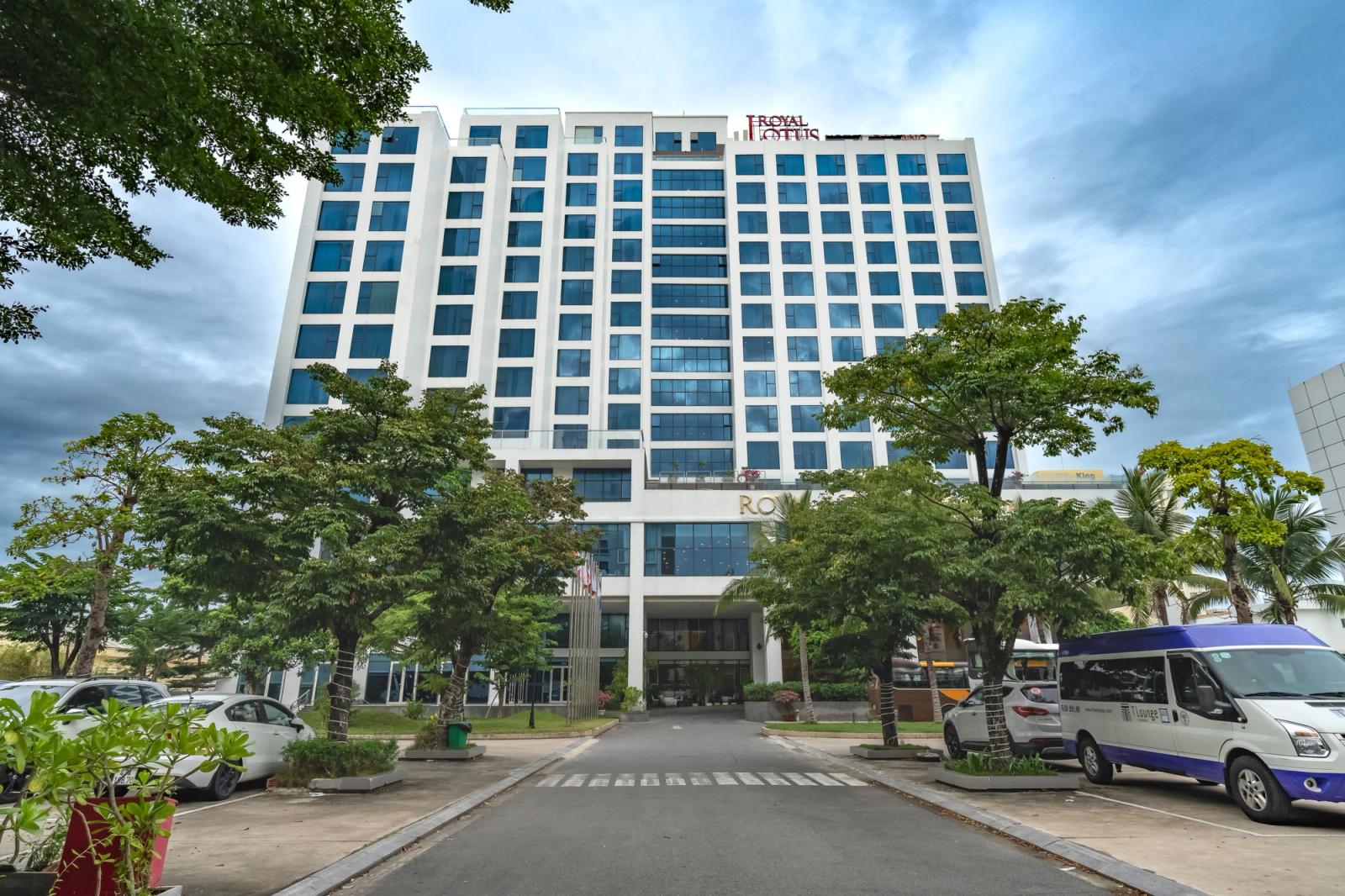 Front view of a modern hotel building surrounded by trees and parked vehicles under a cloudy sky