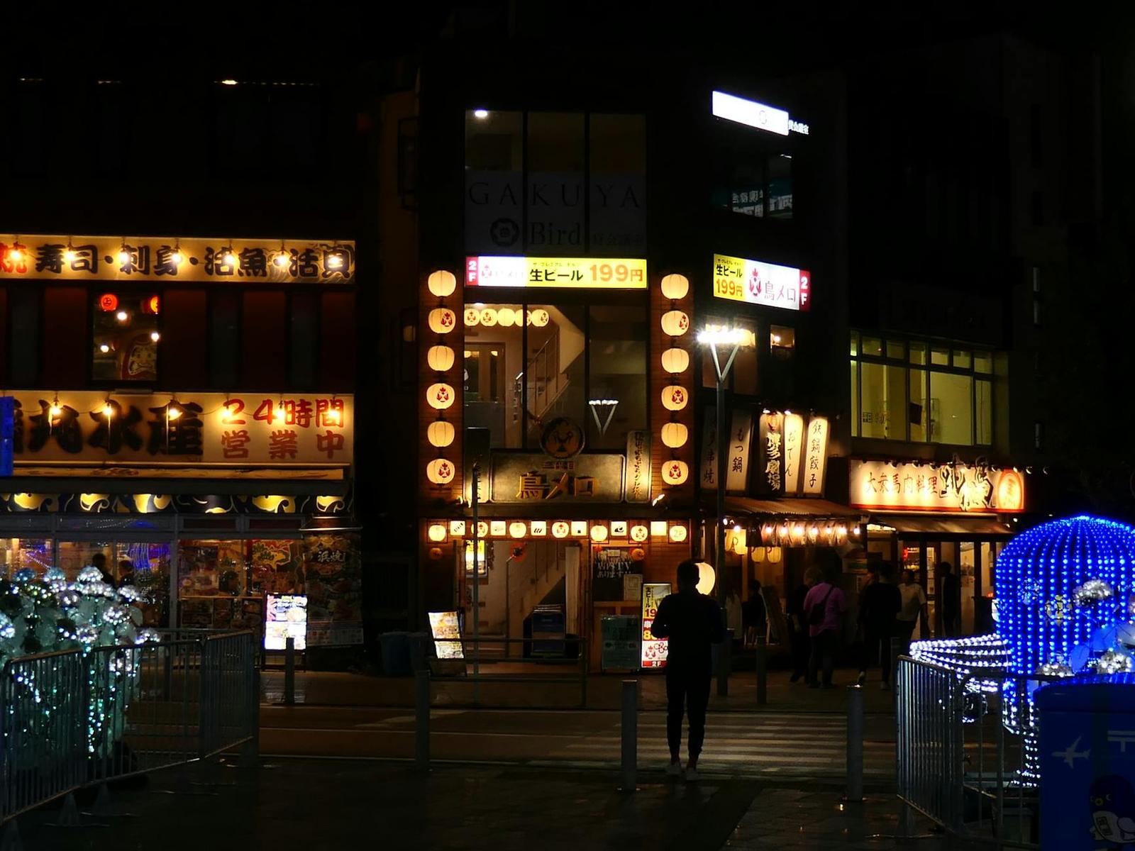 A lone figure crossing a plaza lined with illuminated shopfronts at night