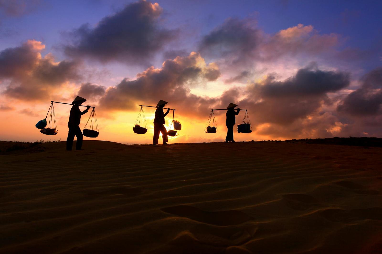 Silhouetted figures carrying shoulder baskets across sand dunes under a fiery sunset sky