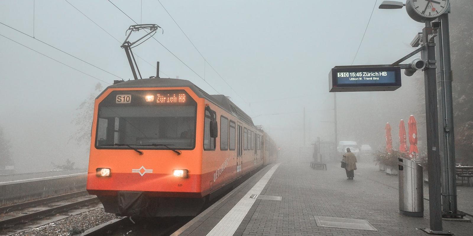 An orange commuter train waiting at a foggy platform beside a lone passenger